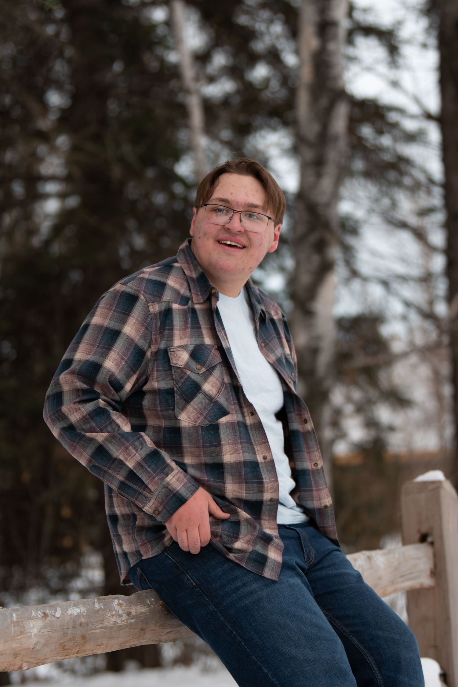 A person in a plaid flannel jacket and jeans sits casually on a wooden fence while smiling outdoors.