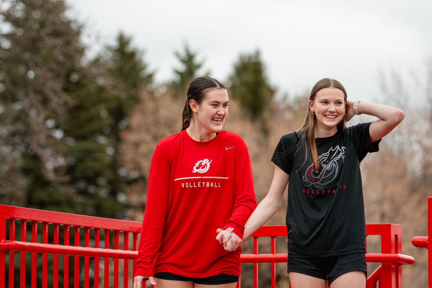 Two young women in athletic wear smile and hold hands on a red bridge outdoors.