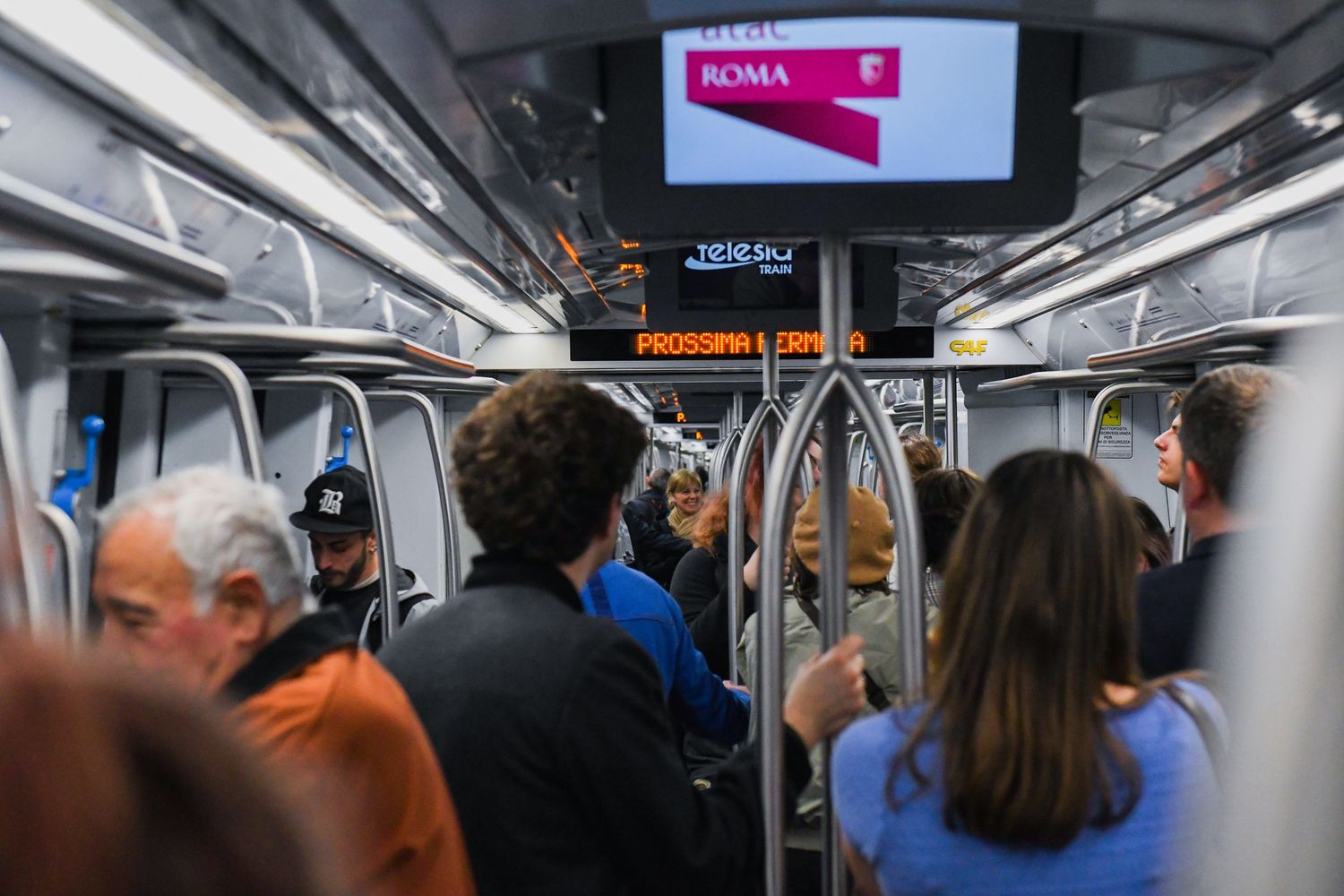 Crowded subway car interior with digital display screen showing transit information.