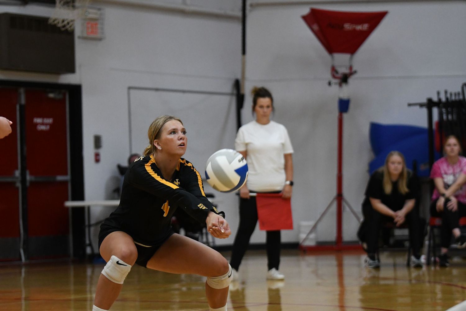 Volleyball player in black and gold uniform crouches to receive serve during match.