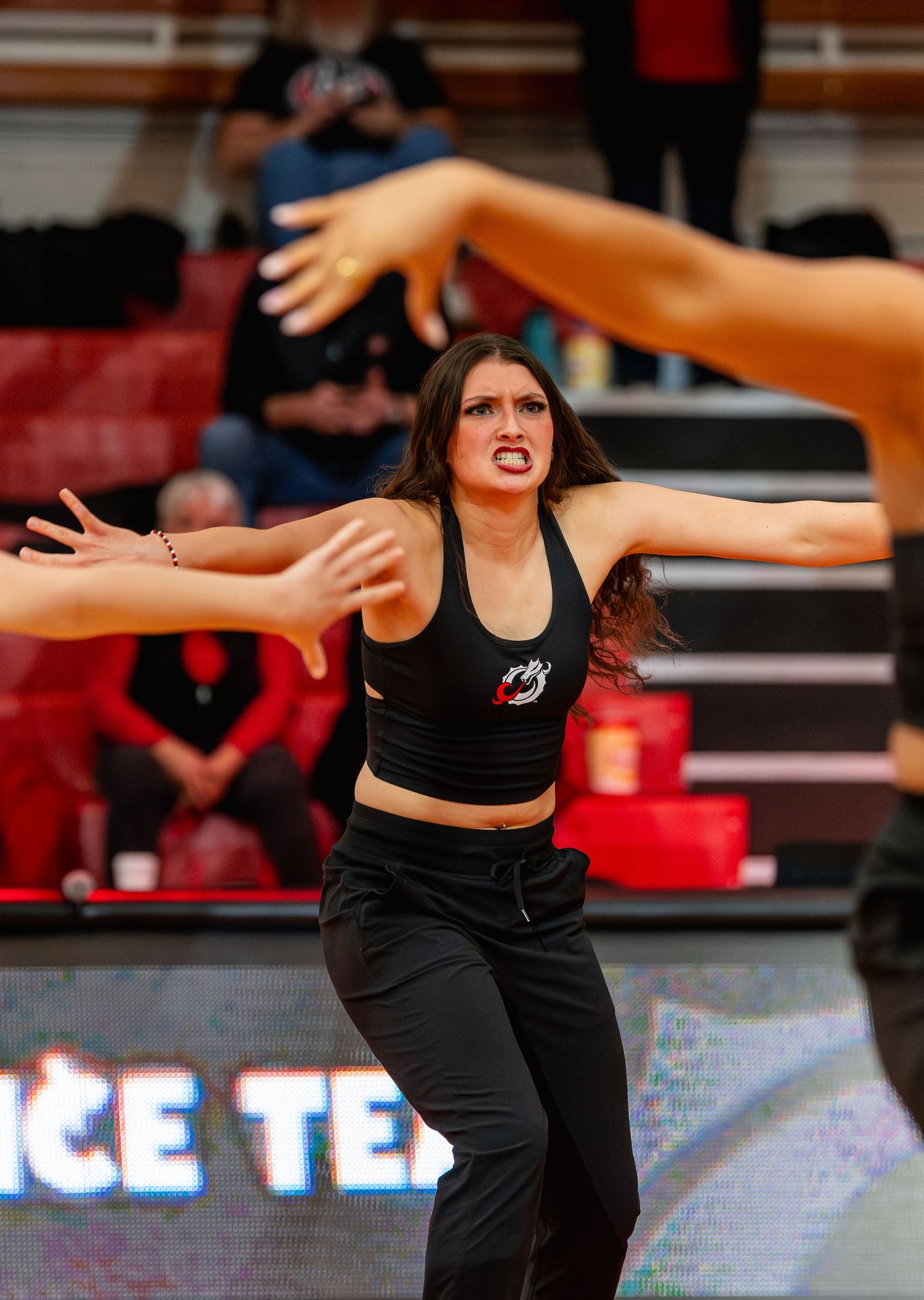 A dancer in black athletic wear performs an expressive movement with outstretched arms on a basketball court.