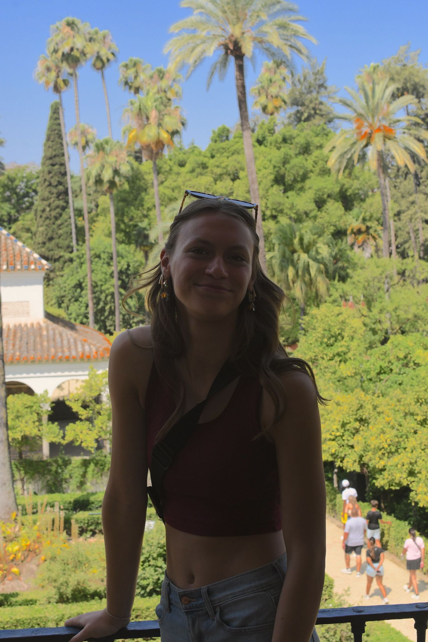 Tourist posing on balcony overlooking lush garden with palm trees and Mediterranean architecture in background.