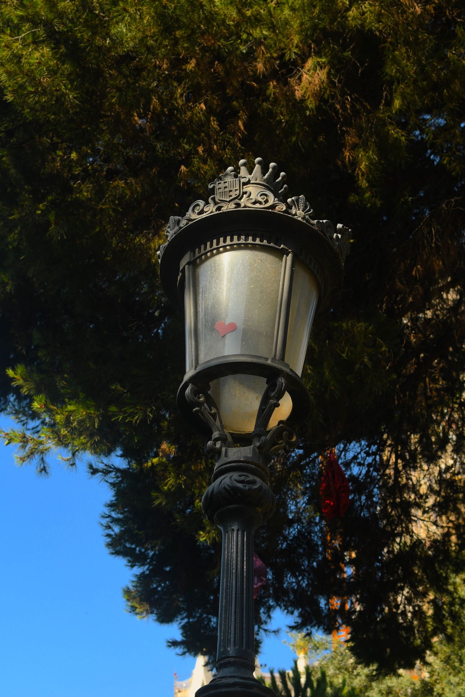 Ornate vintage street lamp with decorative metalwork against blue sky and tree branches.