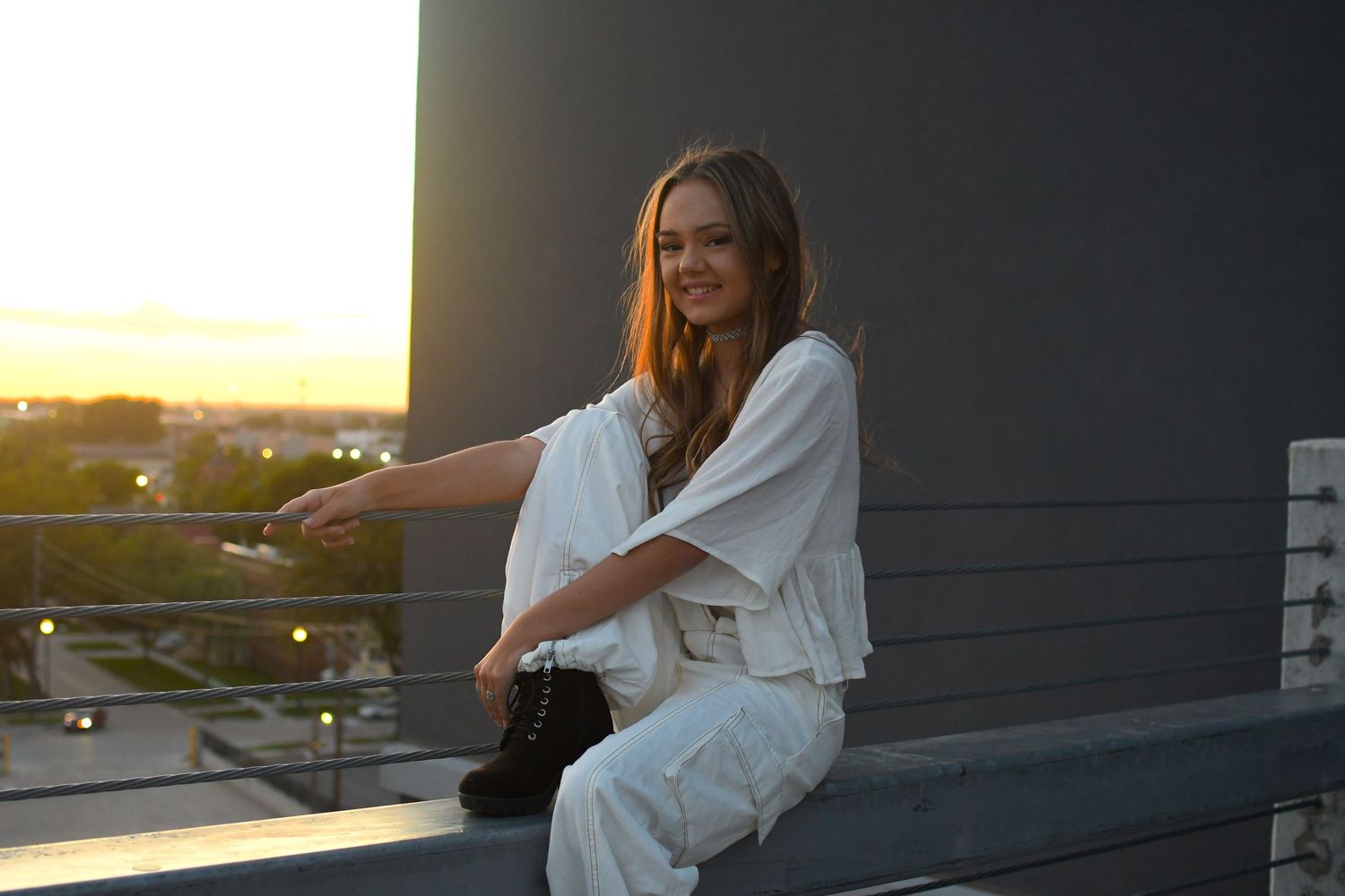 Person in white robe relaxing on balcony during golden sunset overlooking cityscape.