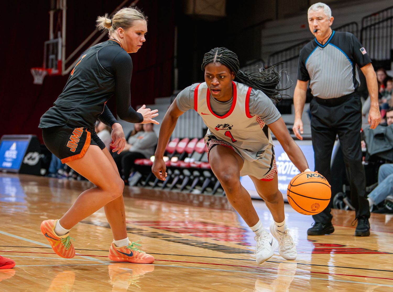 Basketball player dribbles past defender during intense game action on indoor court with referee watching.