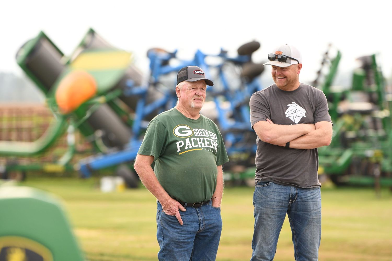 Farmers stand in a field discussing equipment while examining green agricultural machinery in the background.