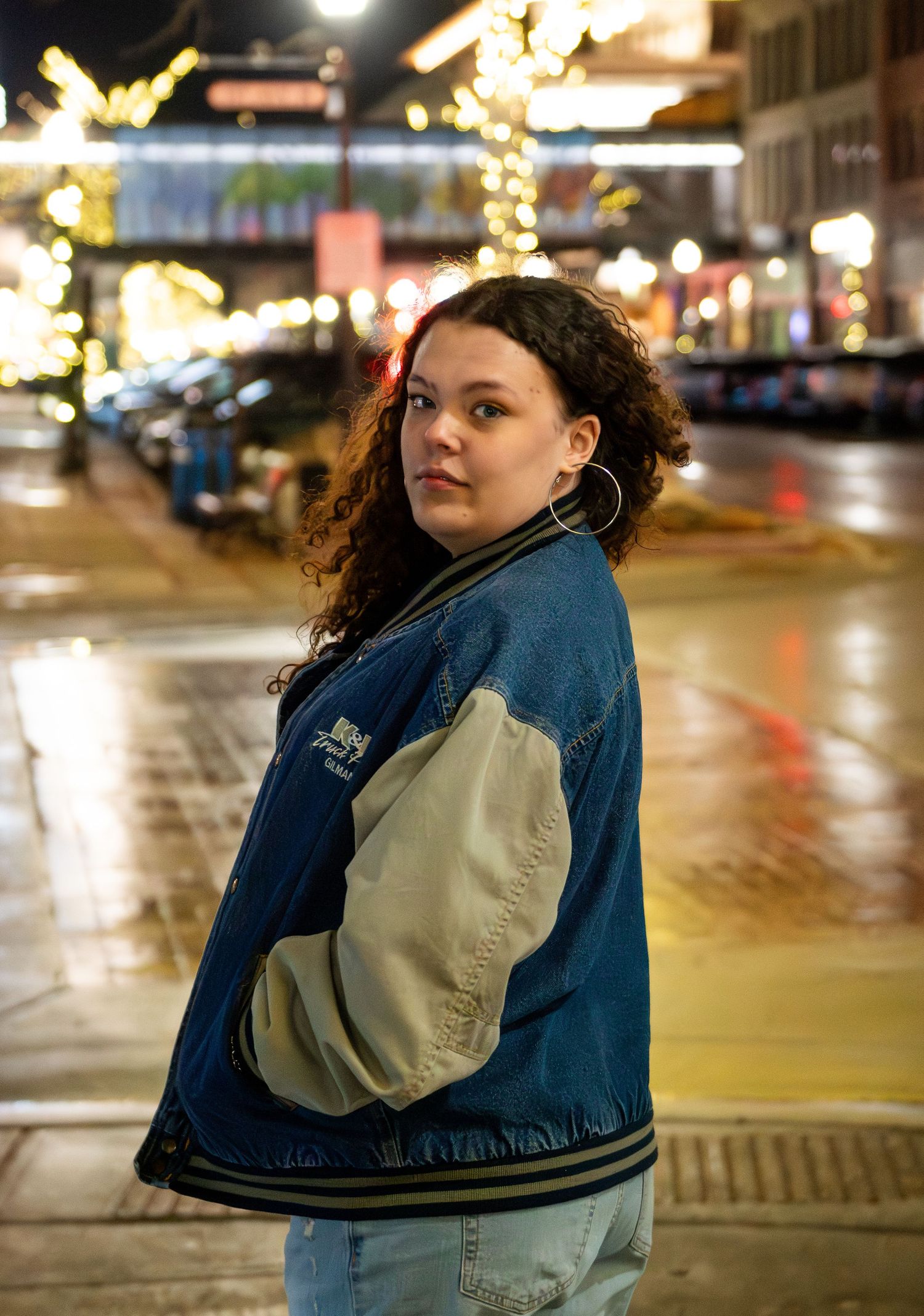 A person in a varsity jacket stands on a wet city street at night with holiday lights glowing in the background.