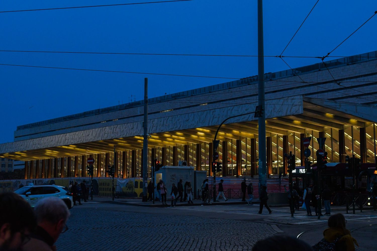 Modern illuminated train station exterior at dusk with people waiting under covered platform.