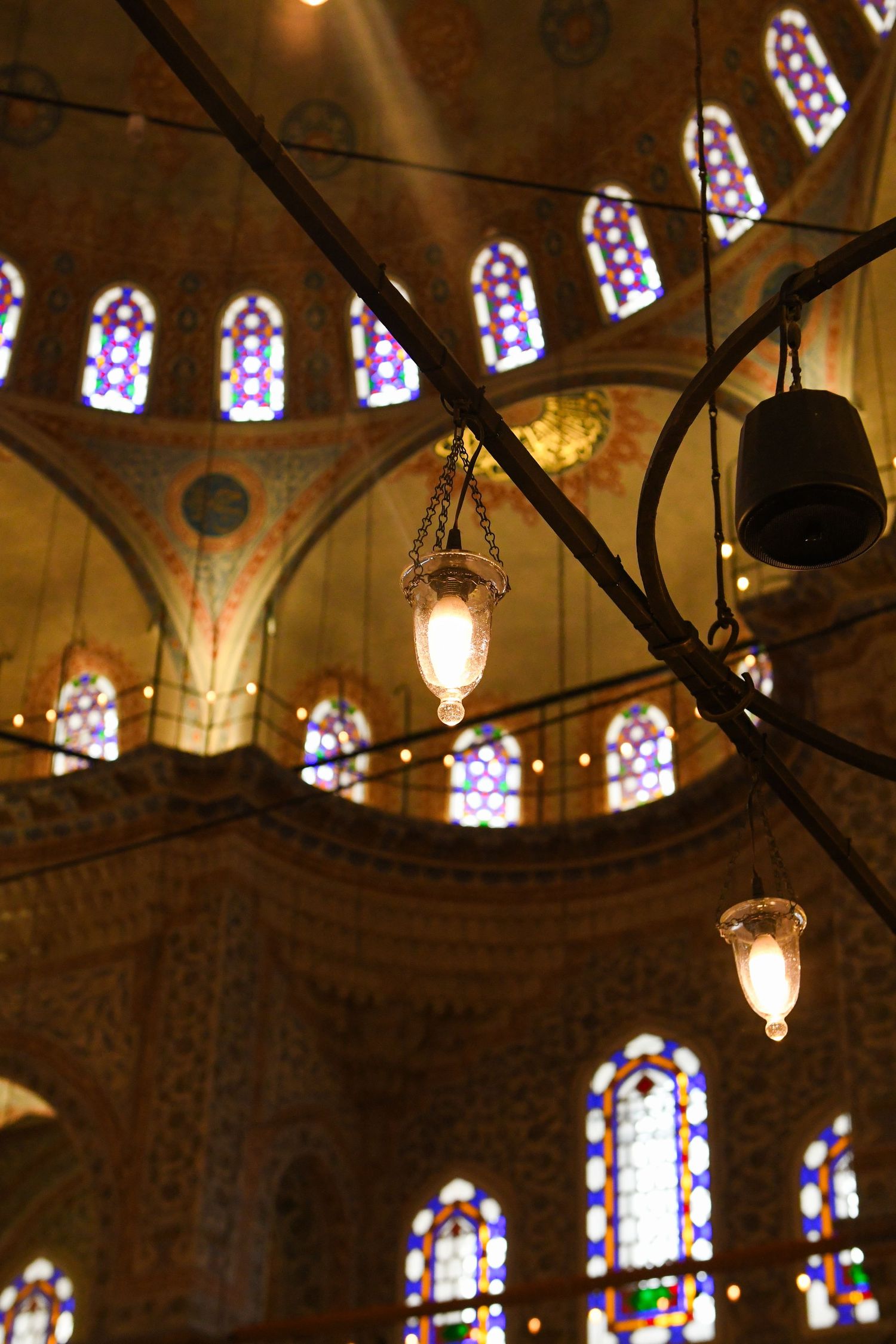 Ornate stained glass windows and hanging lanterns illuminate the interior of a historic mosque or cathedral.