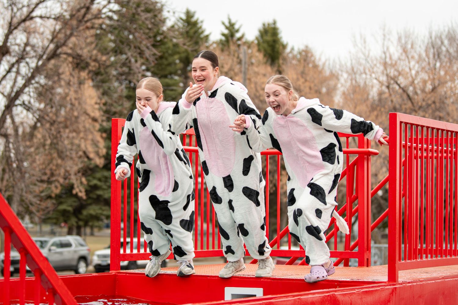 Three girls wearing matching cow print onesie costumes pose playfully on a red platform outdoors.