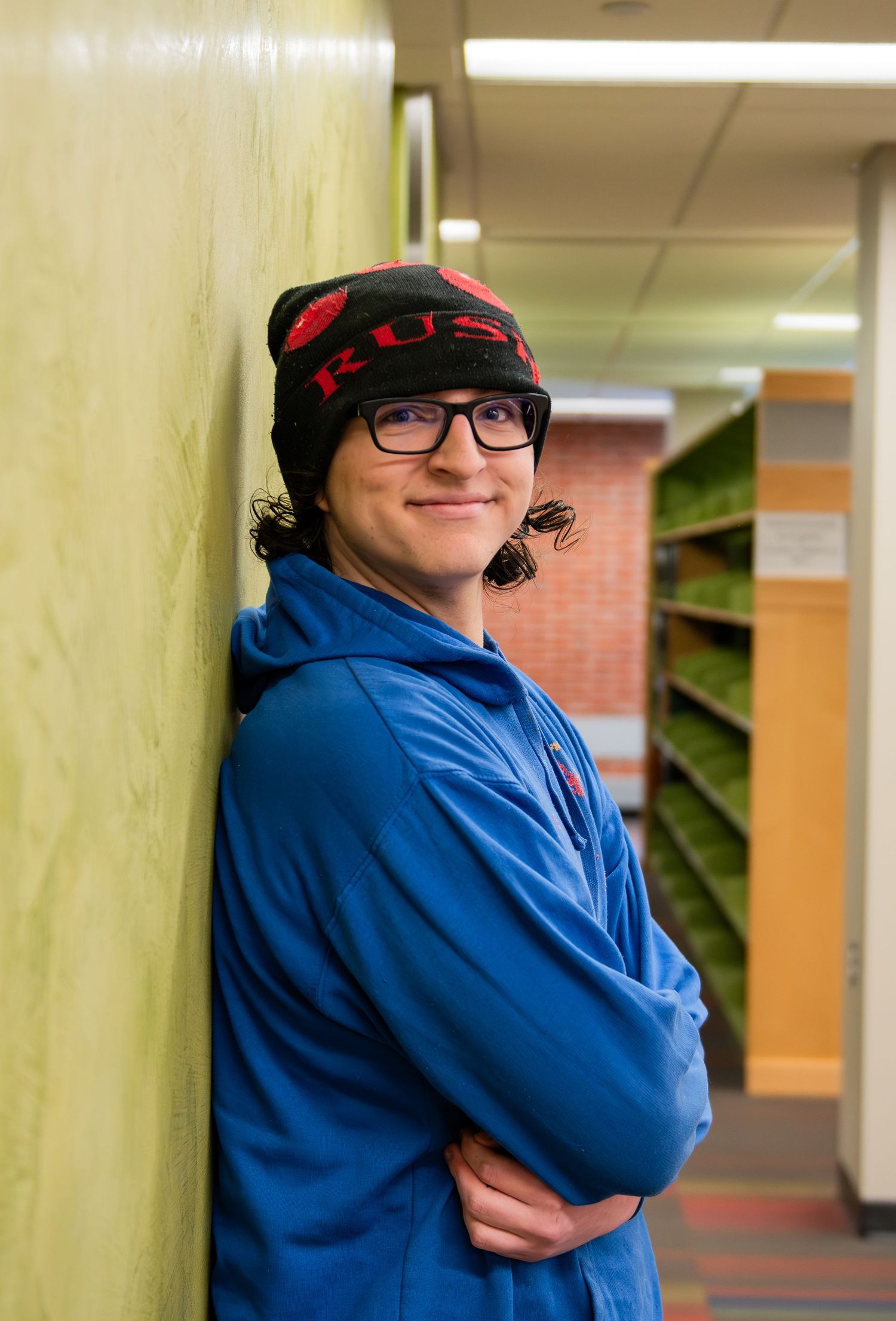 Student in blue hoodie and winter hat smiles while standing against green pillars in school hallway.