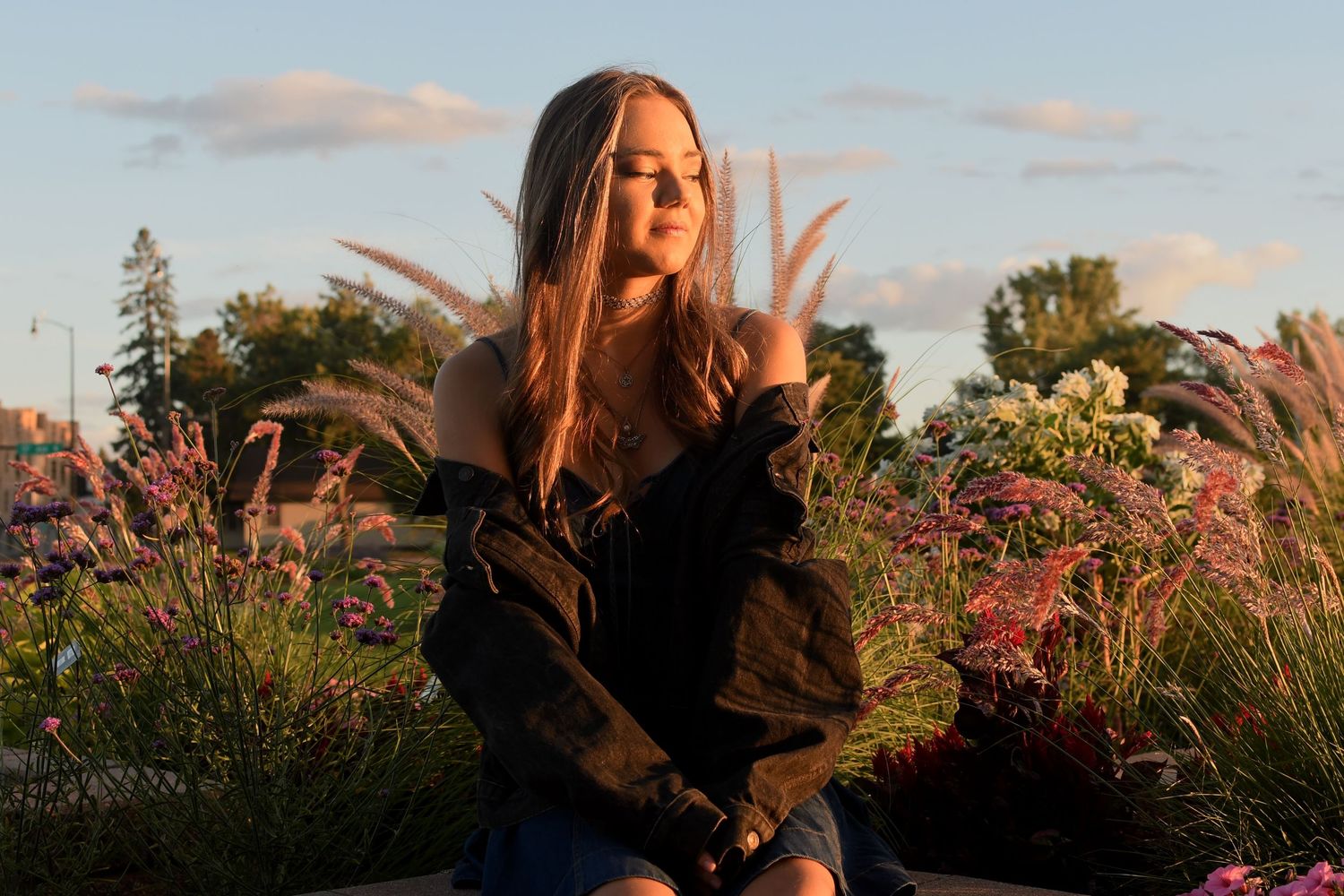 A person in a brown jacket sits in a field of wildflowers during golden hour sunset.