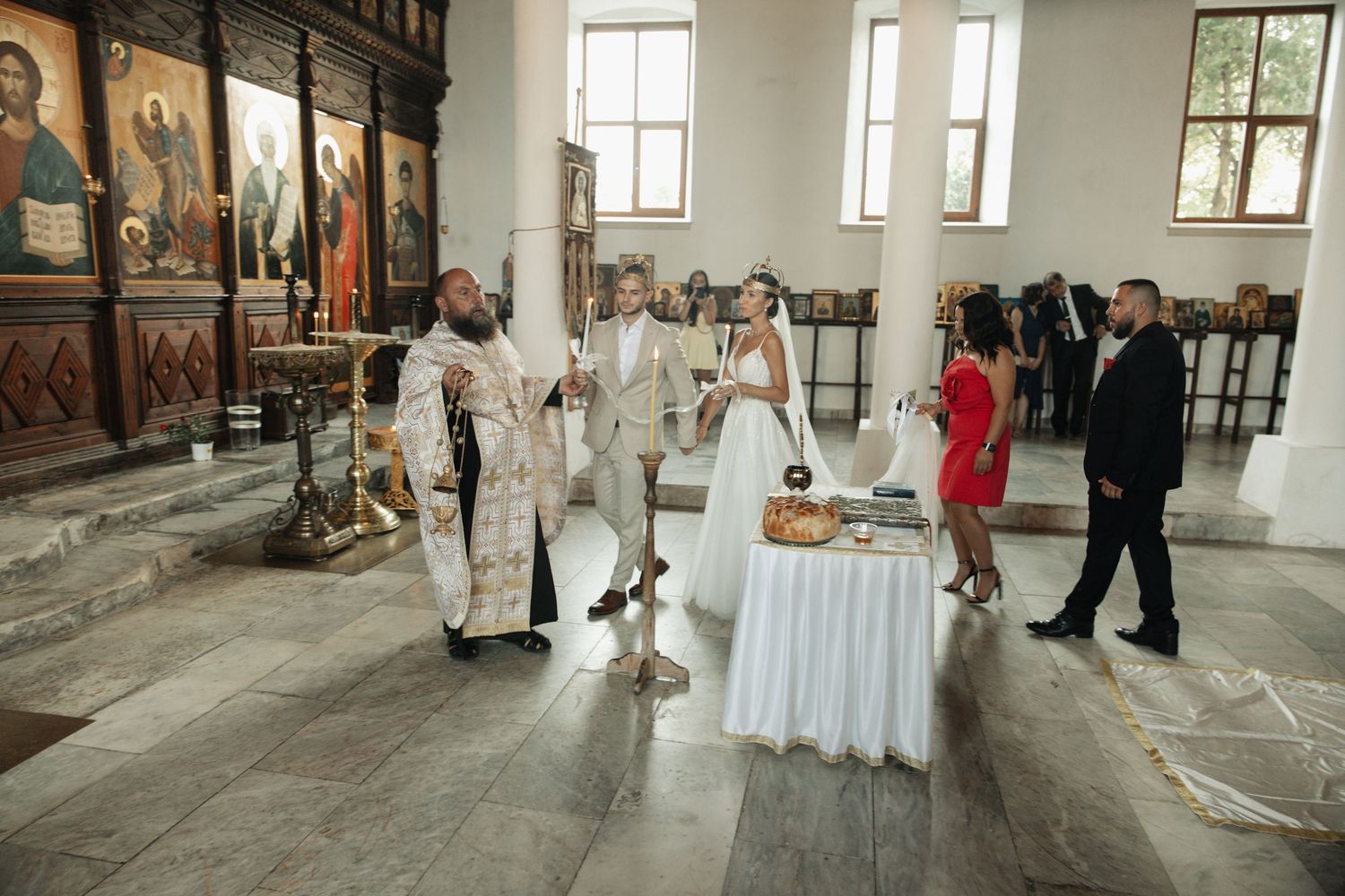 Orthodox wedding ceremony inside a church with priest, bride in white gown, and guests gathered around altar.