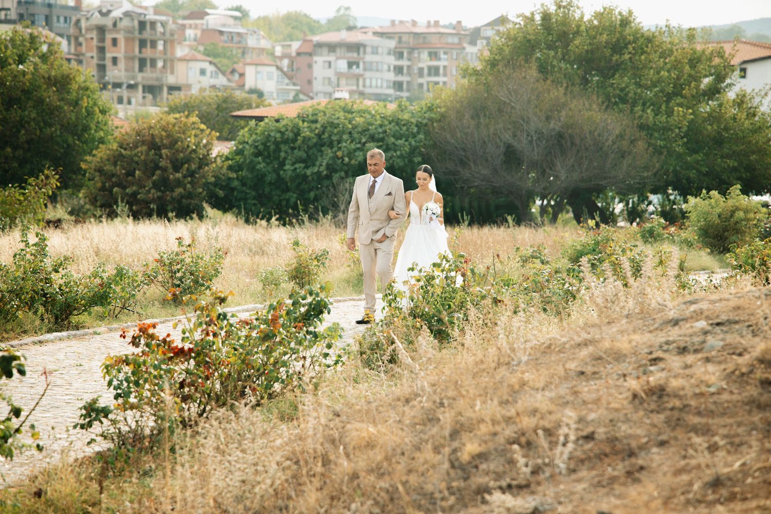 Bride and groom walking hand in hand along a garden path surrounded by wildflowers and greenery on their wedding day.