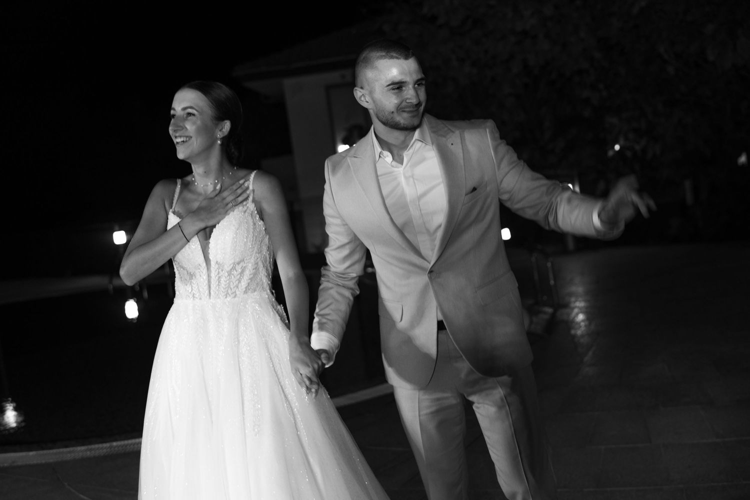 Bride and groom hold hands smiling at elegant outdoor evening wedding reception.