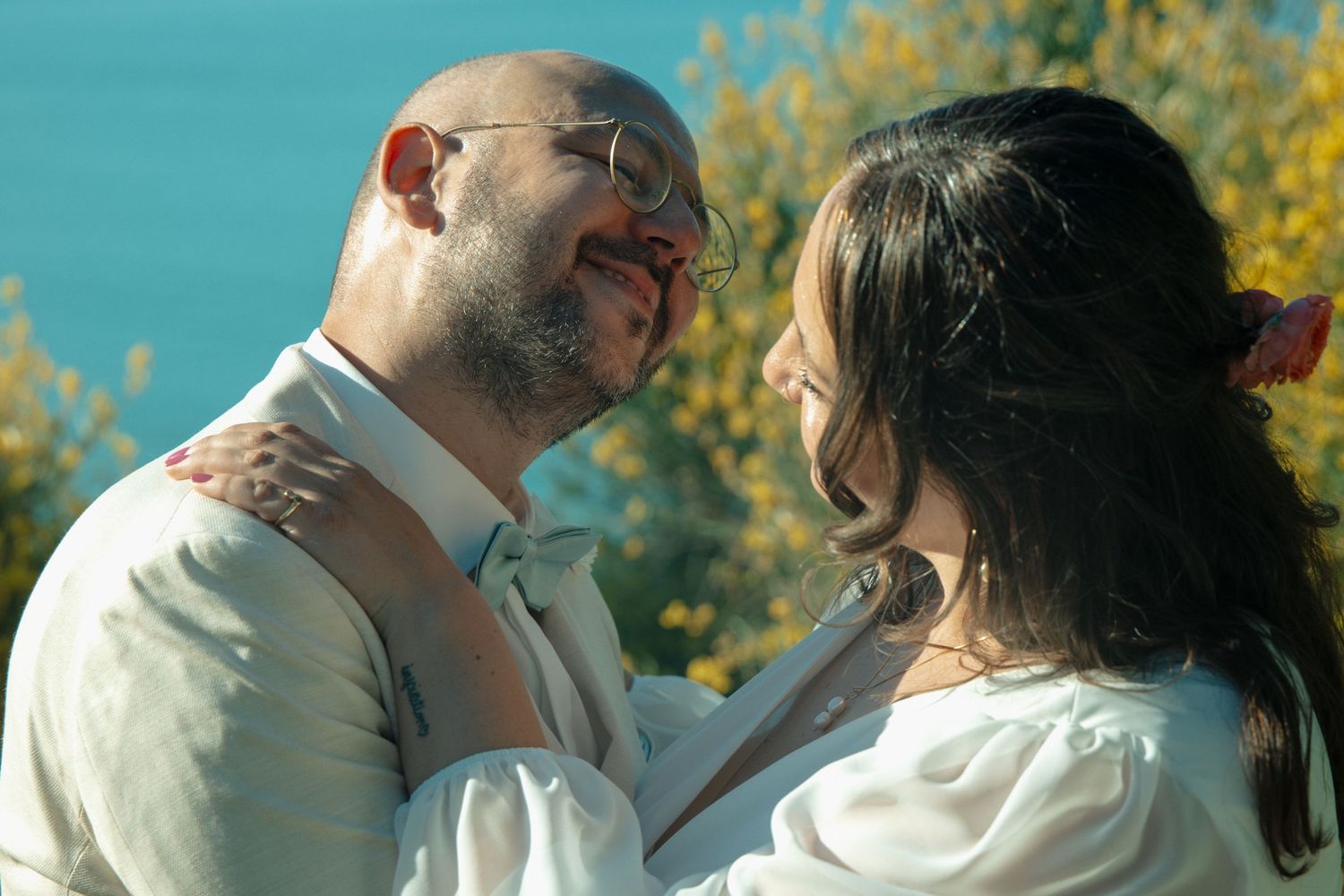 A smiling bald man with glasses and a woman in white embrace outdoors with yellow flowers and blue sea behind them.