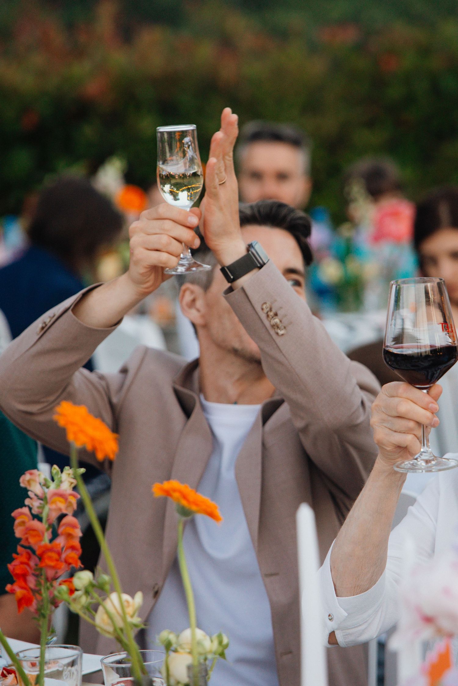 Man in beige blazer raises champagne glass for a toast at an outdoor wedding reception.