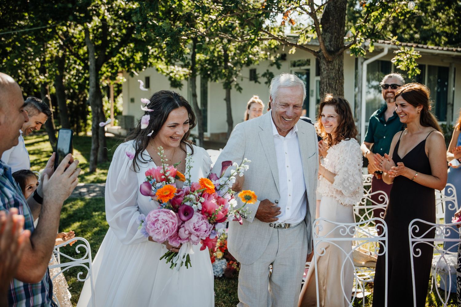 Smiling bride with pink peony bouquet walks with escort past applauding guests at outdoor wedding.