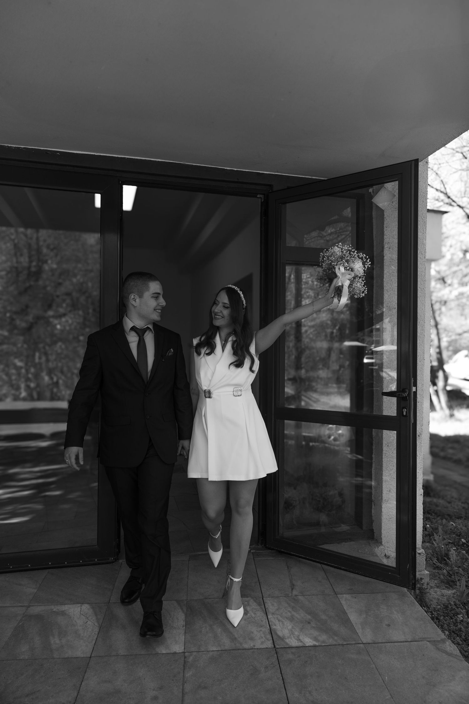 Black and white photo of a bride in short white dress holding bouquet and groom in suit walking out of building.