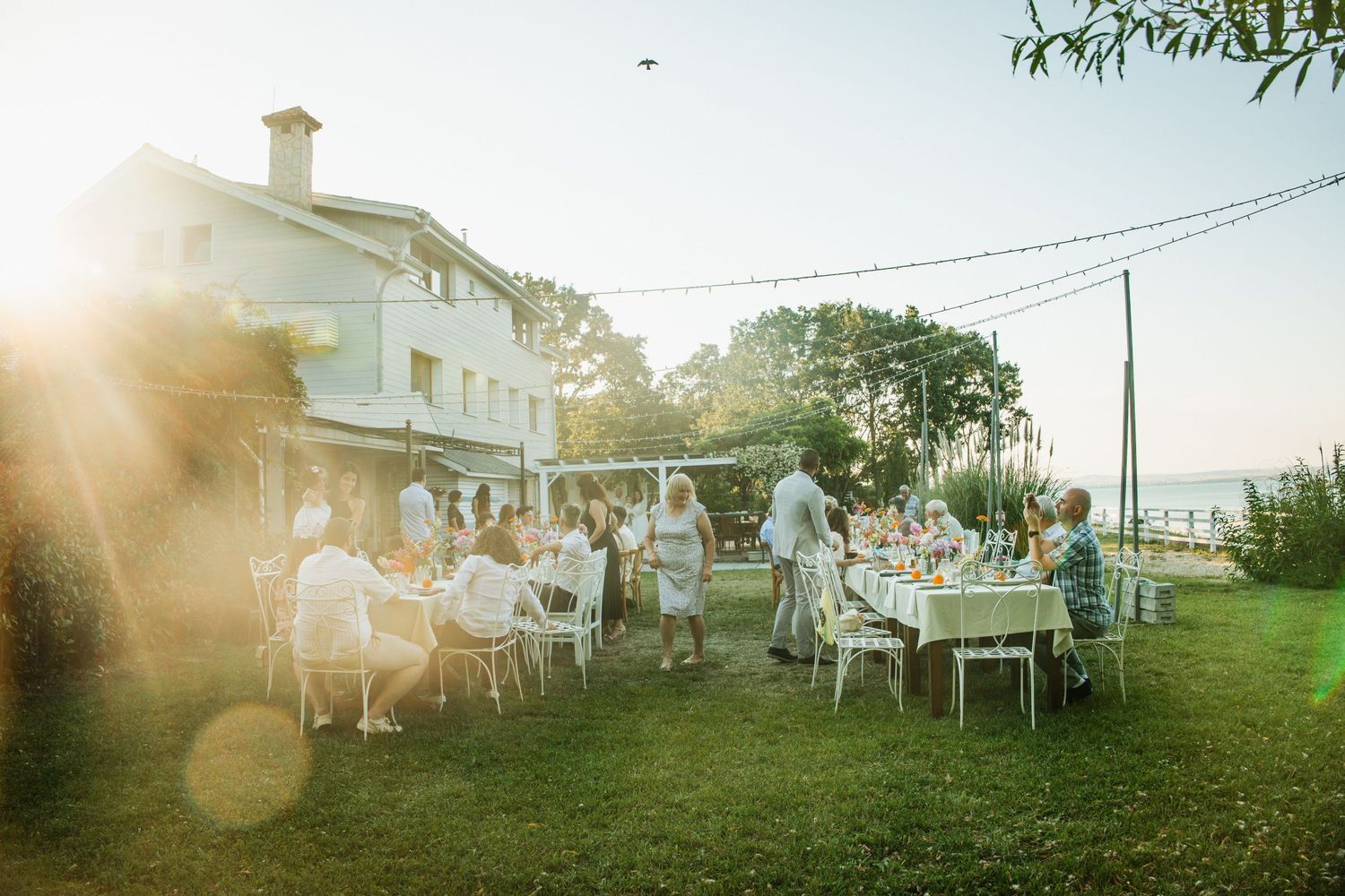 Outdoor garden party with guests dining at white tables beside a white house at golden hour sunset.