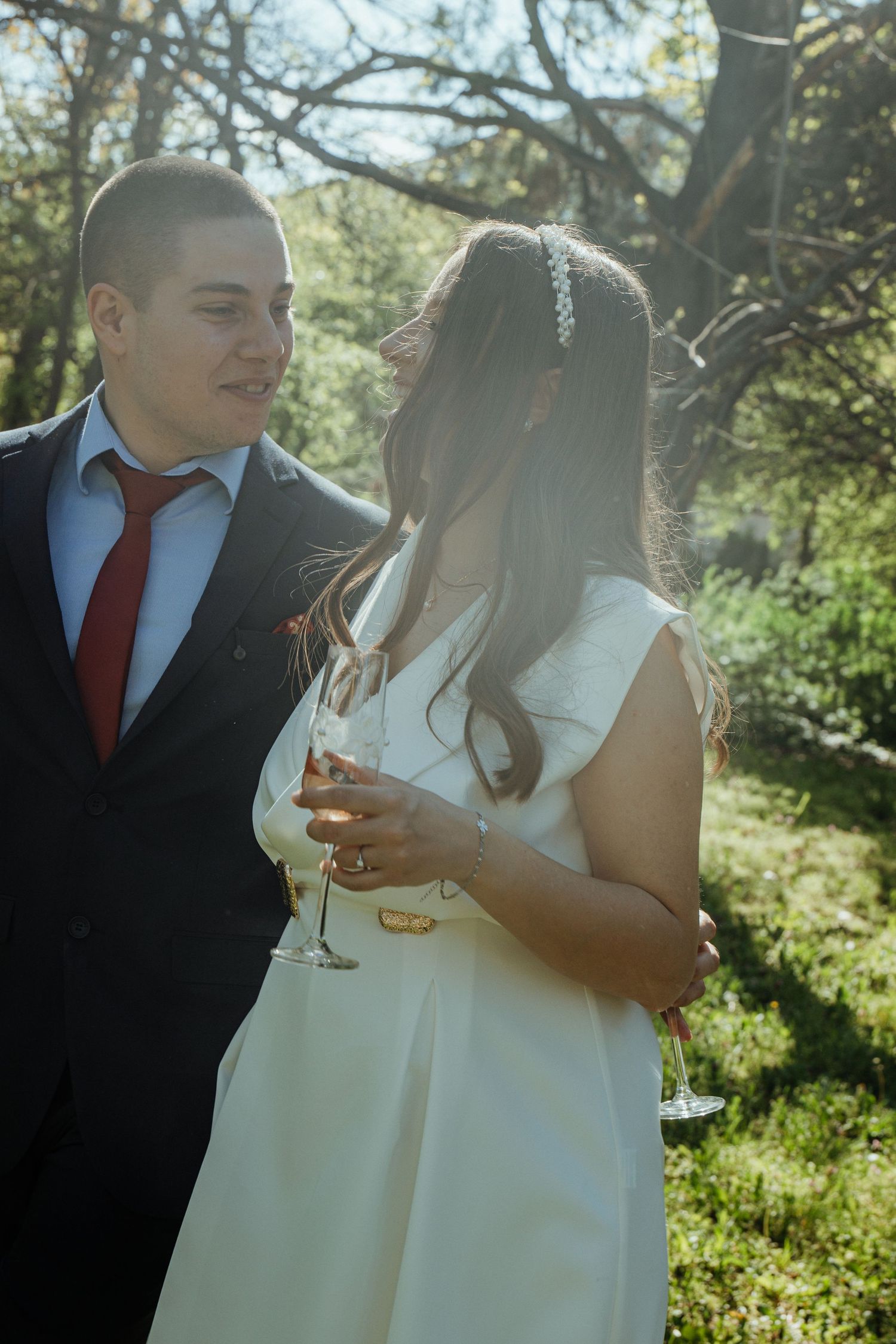 Bride in white dress holding champagne glass stands with groom in navy suit and red tie in a sunlit garden.