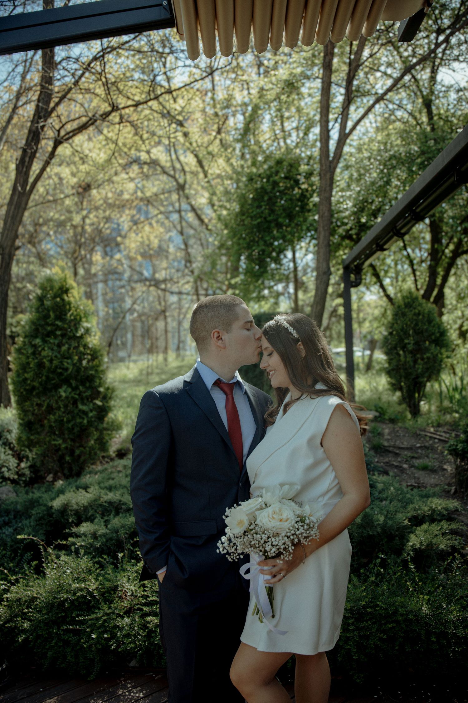 A couple shares a romantic kiss outdoors, bride in white mini dress holding bouquet, groom in dark suit with red tie.