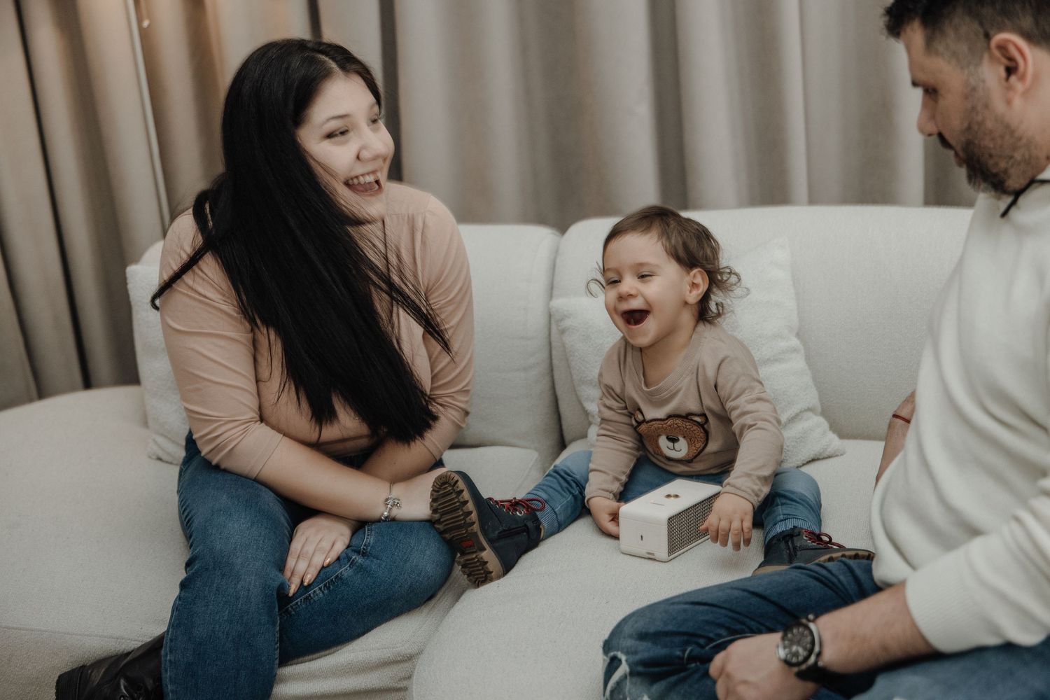 A joyful family of three sits on a white sofa, with a laughing toddler holding a gift box between his parents.
