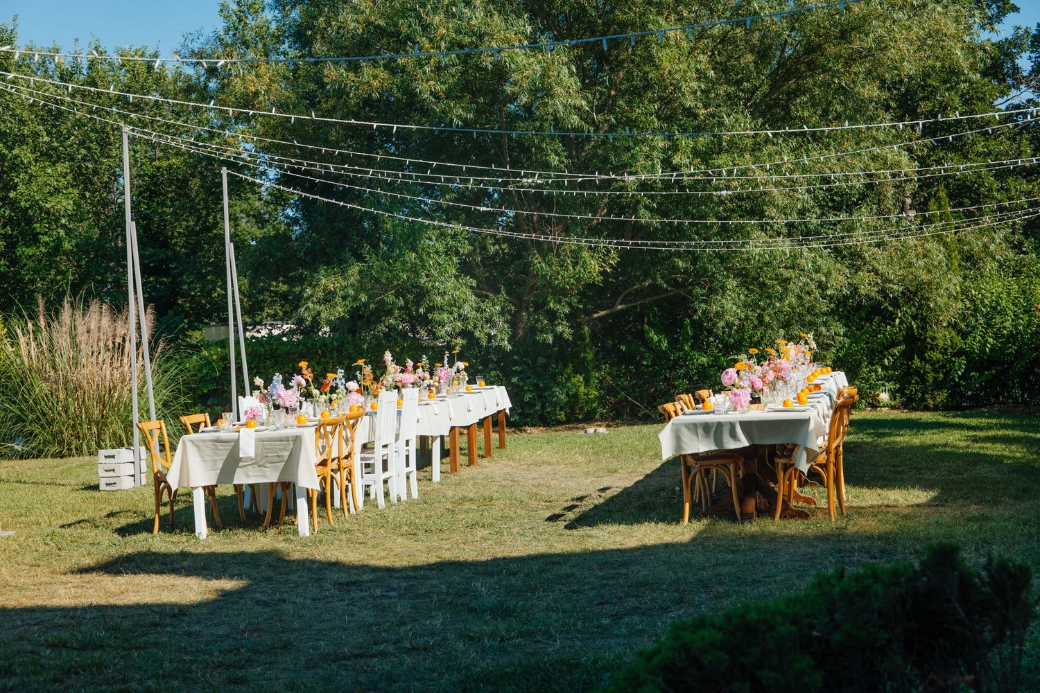 Outdoor wedding reception tables set on green lawn with string lights and colorful floral centerpieces surrounded by trees.
