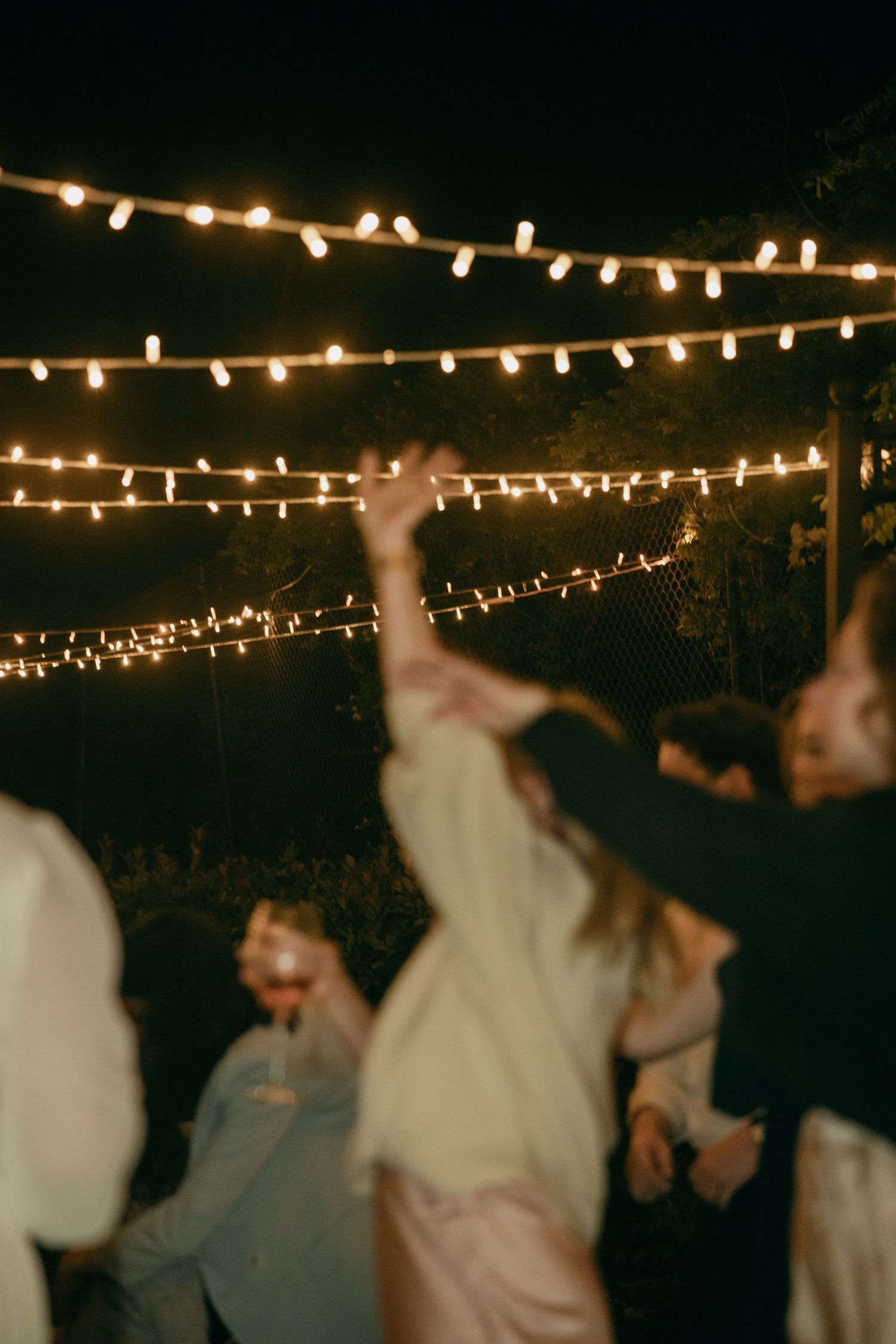 Guests dance under glowing string lights at an outdoor nighttime wedding reception party.