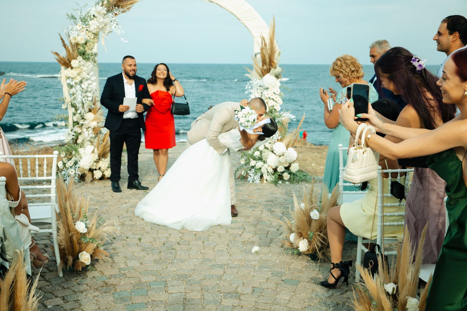 Bride and groom share a romantic dip kiss at a beachside wedding ceremony with floral arch and guests cheering.
