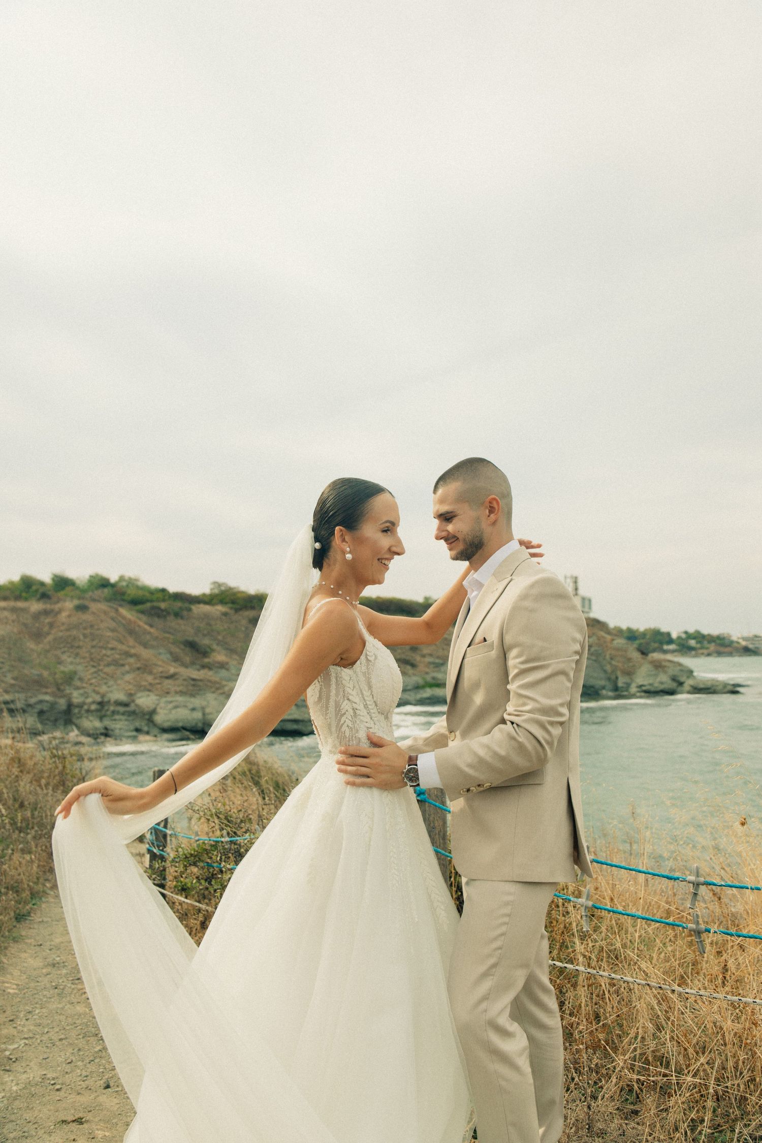 Bride in white gown and groom in beige suit share a romantic moment by the seaside on their wedding day.