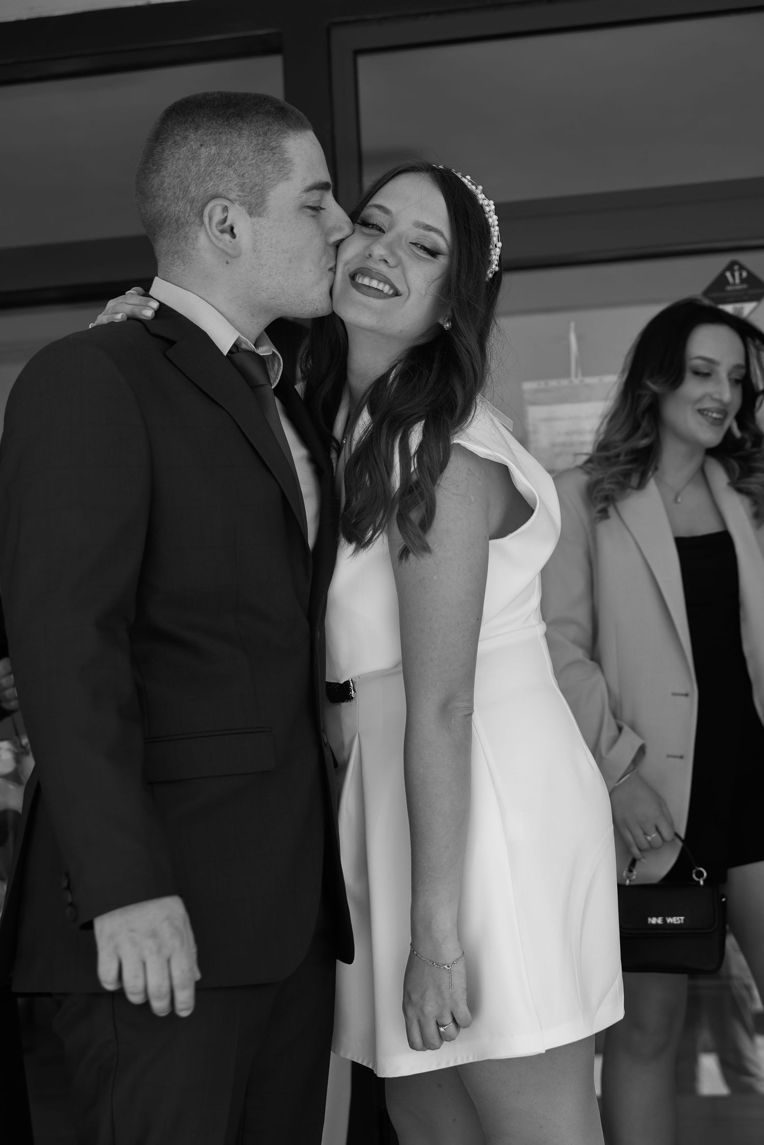 Black and white photo of a groom kissing bride on cheek, bride smiling in white dress with hair accessory.