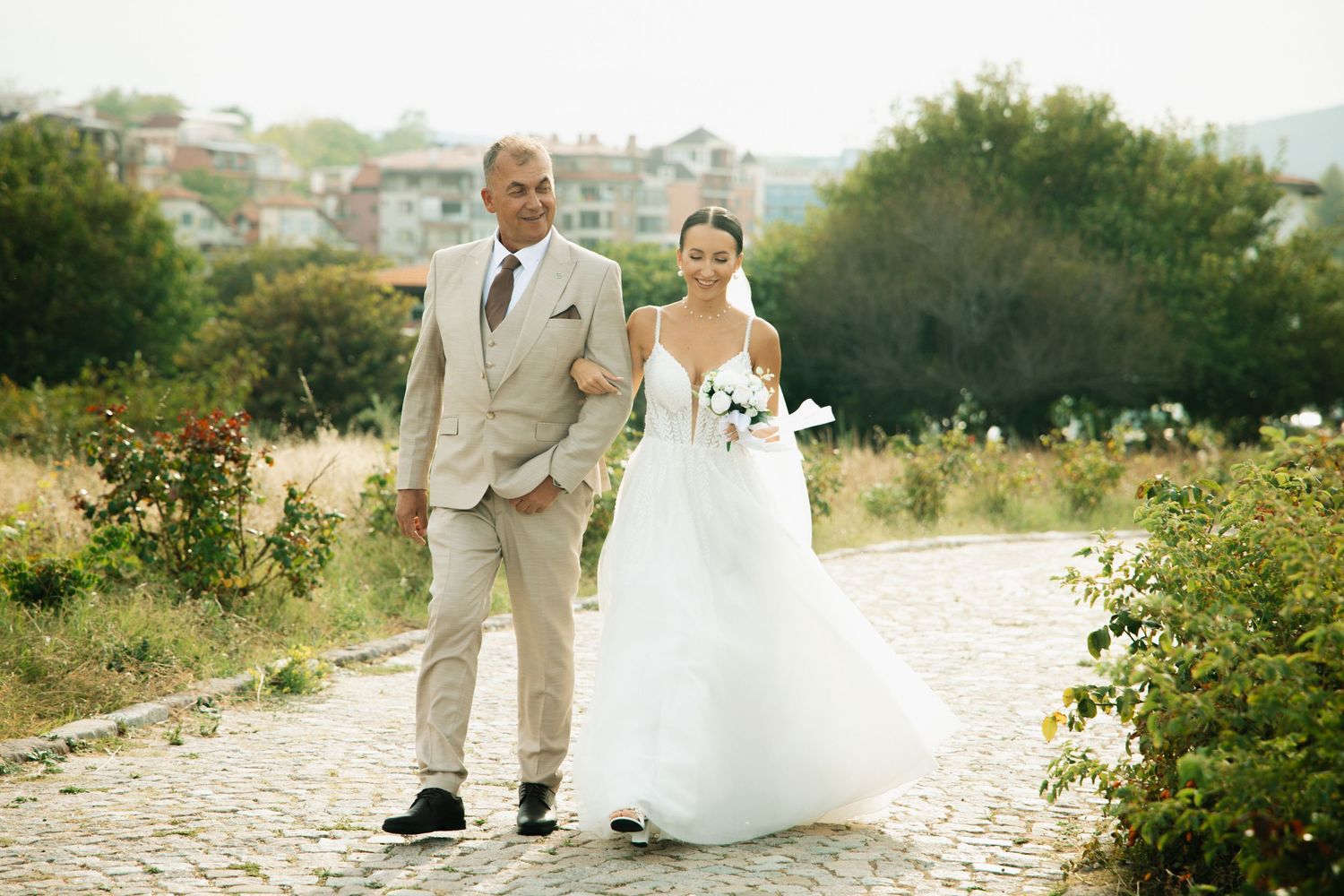 Bride in white gown walks arm-in-arm with man in tan suit on cobblestone path surrounded by greenery.