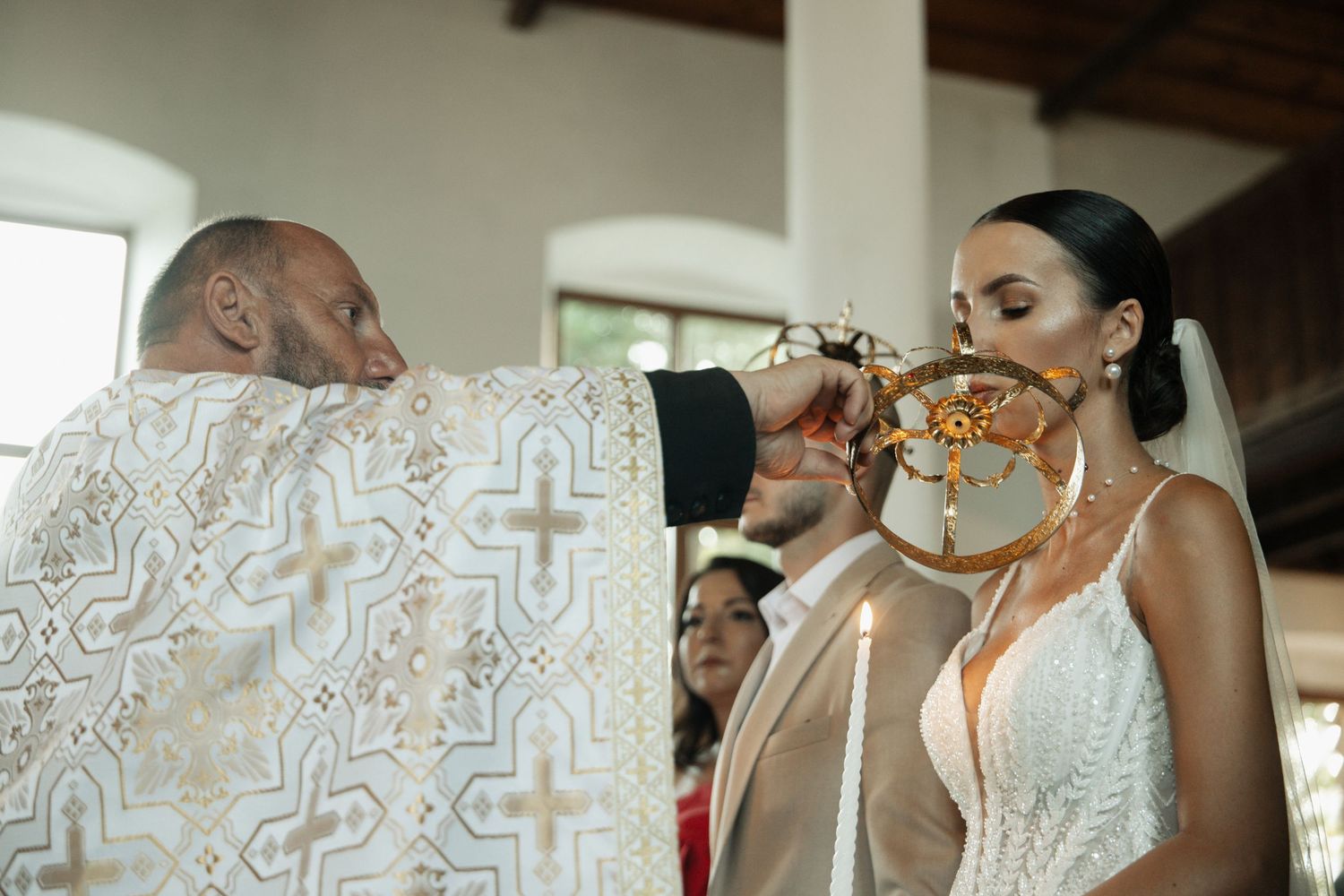 Orthodox priest performing wedding ceremony, offering communion to bride in white gown holding candle in church.