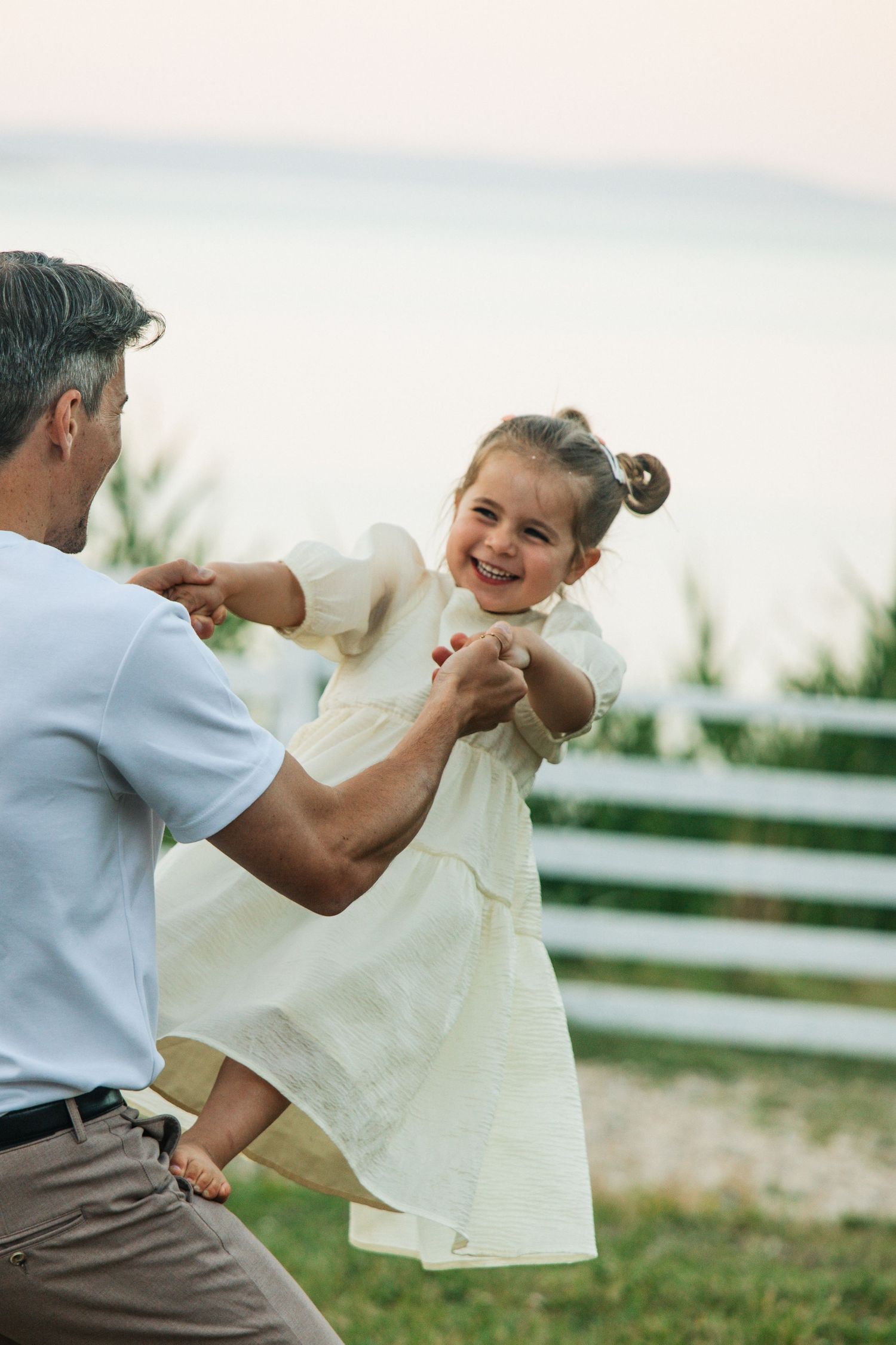 Father lifting smiling little girl in white dress outdoors near white fence with water in background.