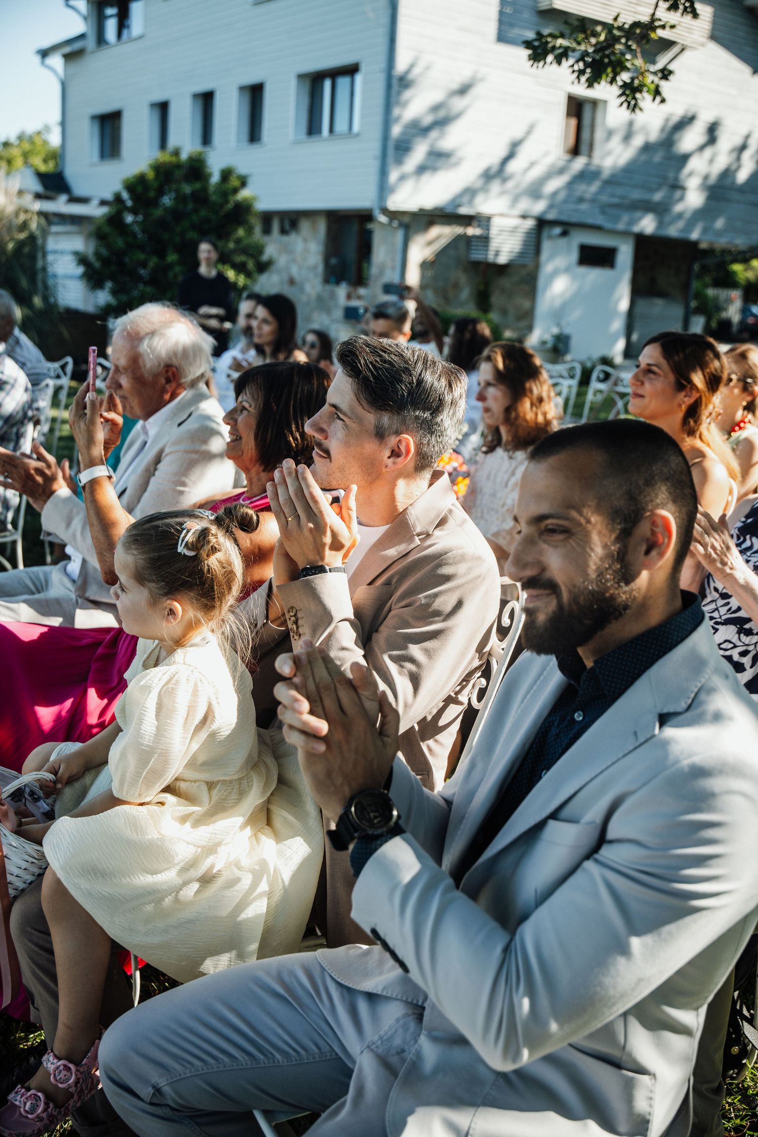 Wedding guests seated outdoors clapping and smiling during a sunny garden ceremony.