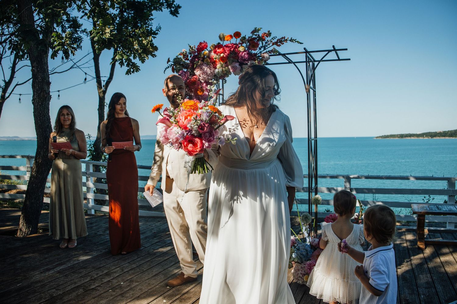 Bride in white gown with floral crown holding bouquet at outdoor seaside wedding ceremony.