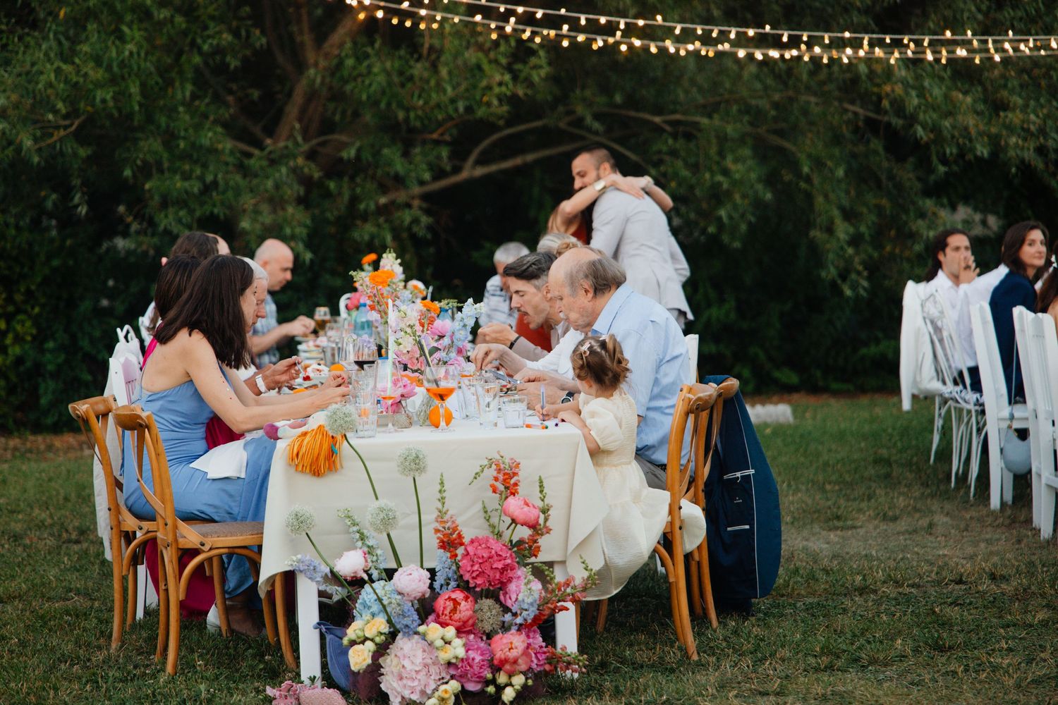 Wedding guests dining at elegantly decorated outdoor table with vibrant floral centerpieces under string lights.