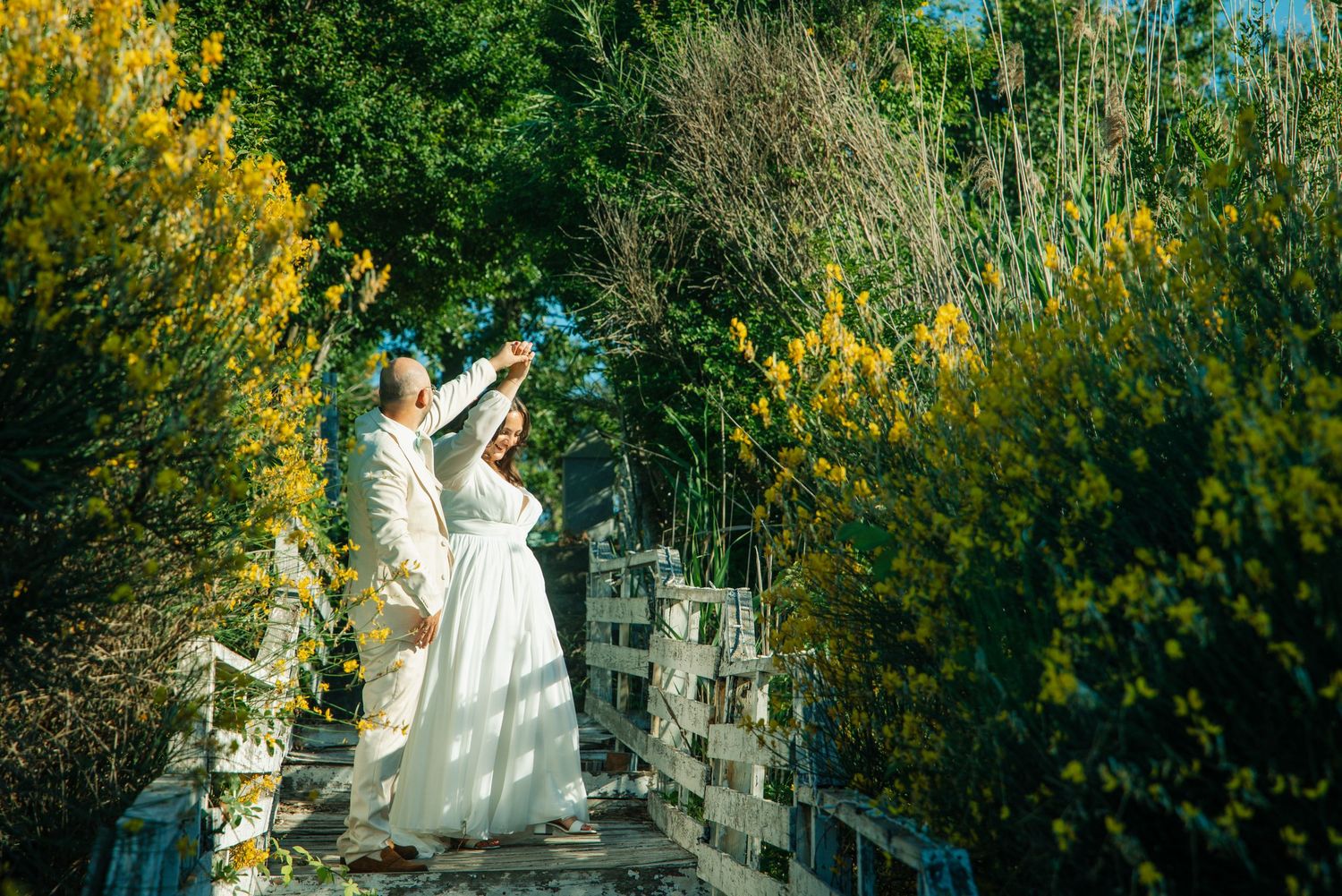 Couple in white attire posing on a garden bridge surrounded by lush green and yellow foliage.