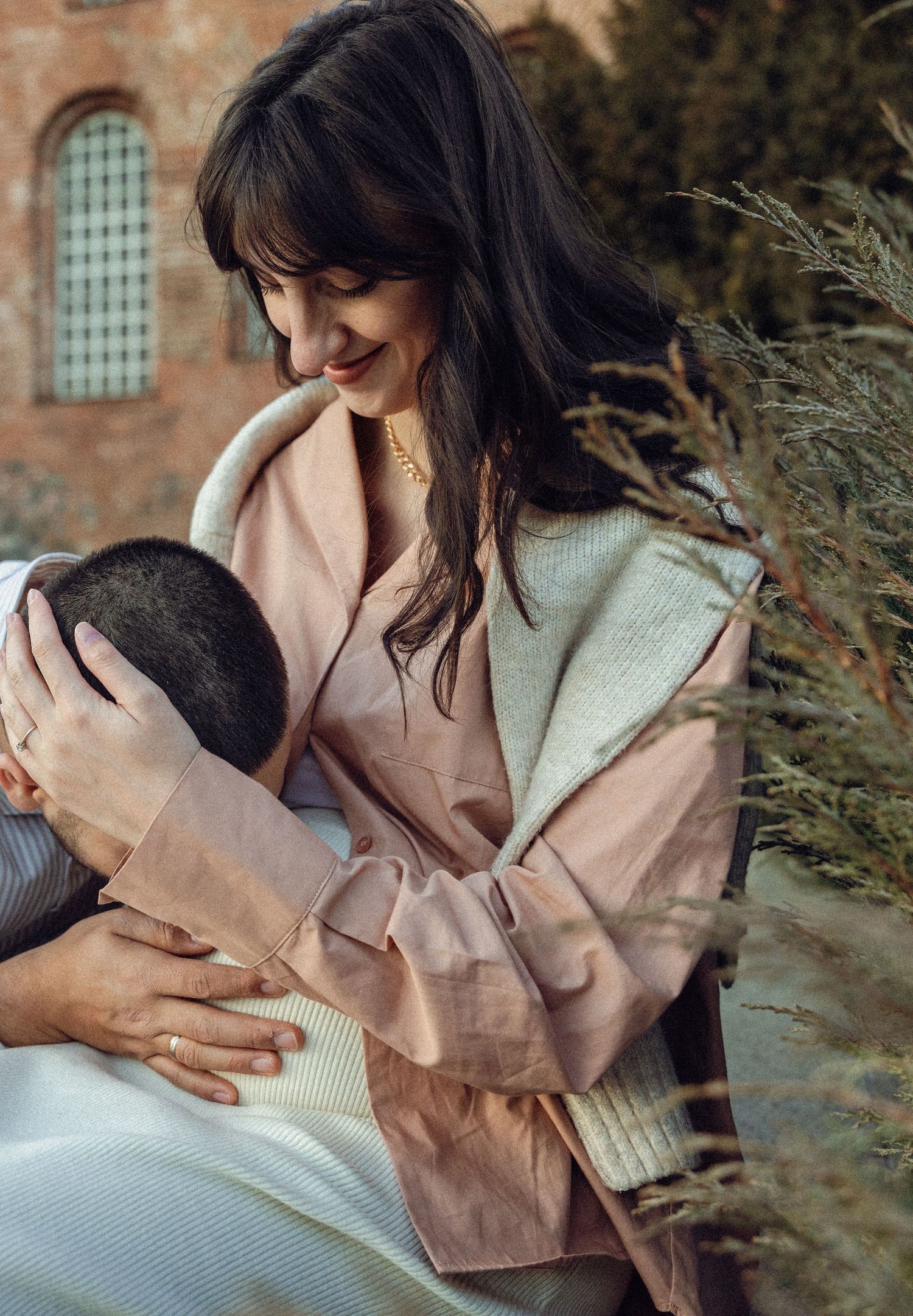 A couple in romantic embrace wearing beige and cream colored clothing against a rustic brick wall background.