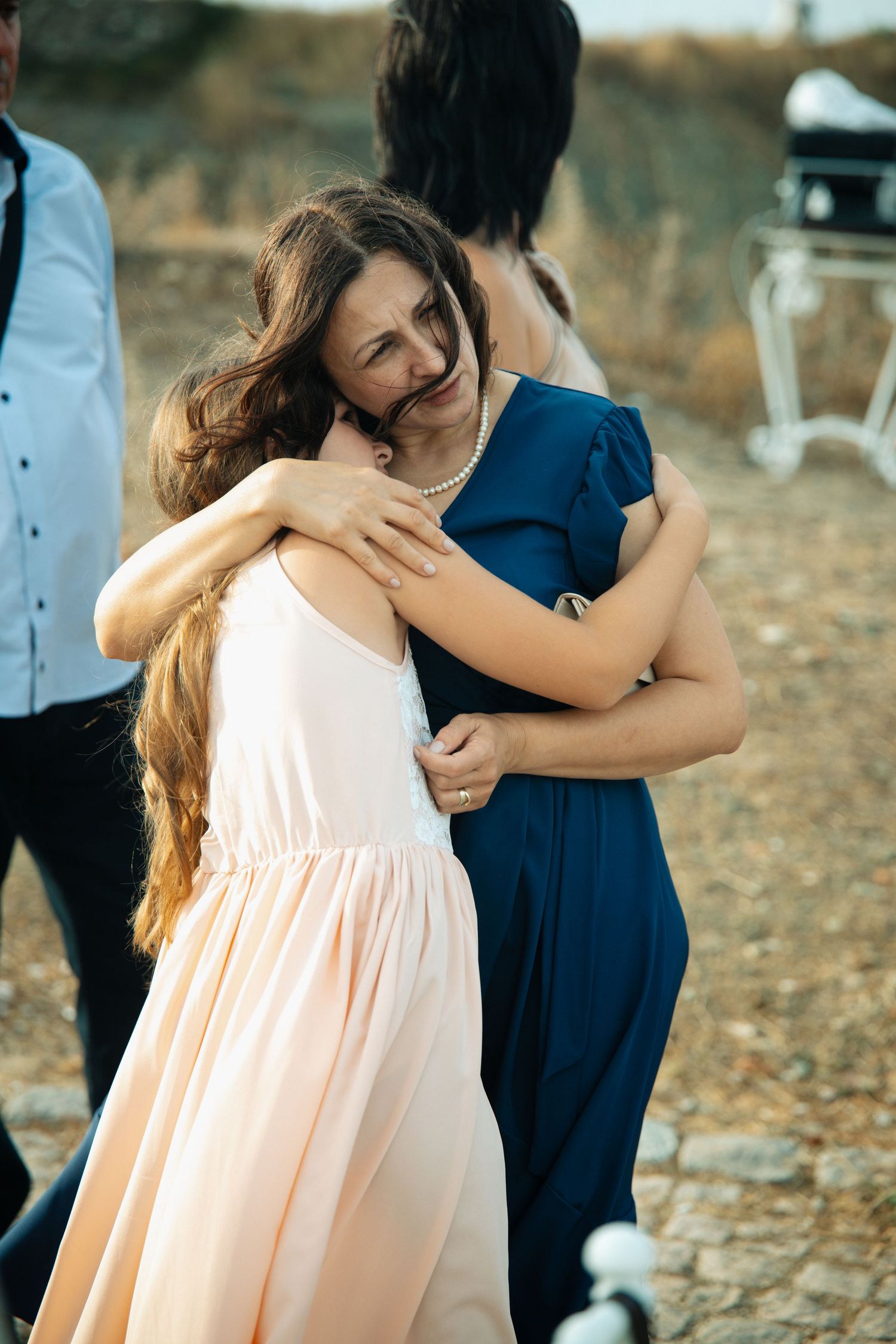 Two women in elegant dresses share an emotional hug at an outdoor wedding celebration.