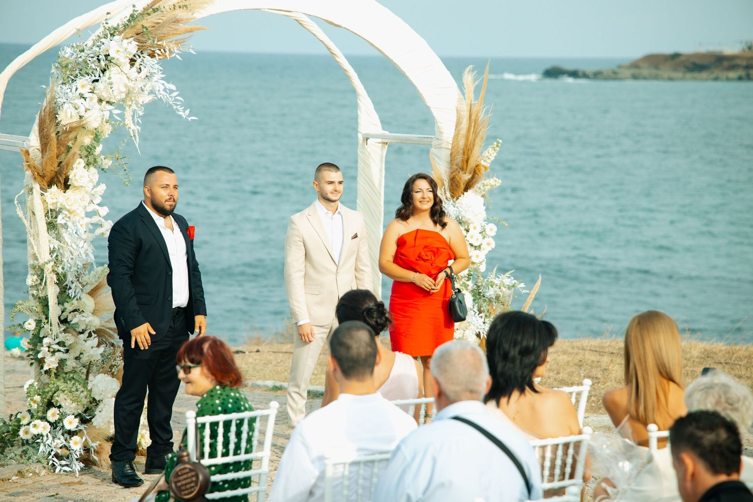 Outdoor beach wedding ceremony with couple and officiant under white floral arch, guests seated by the sea.