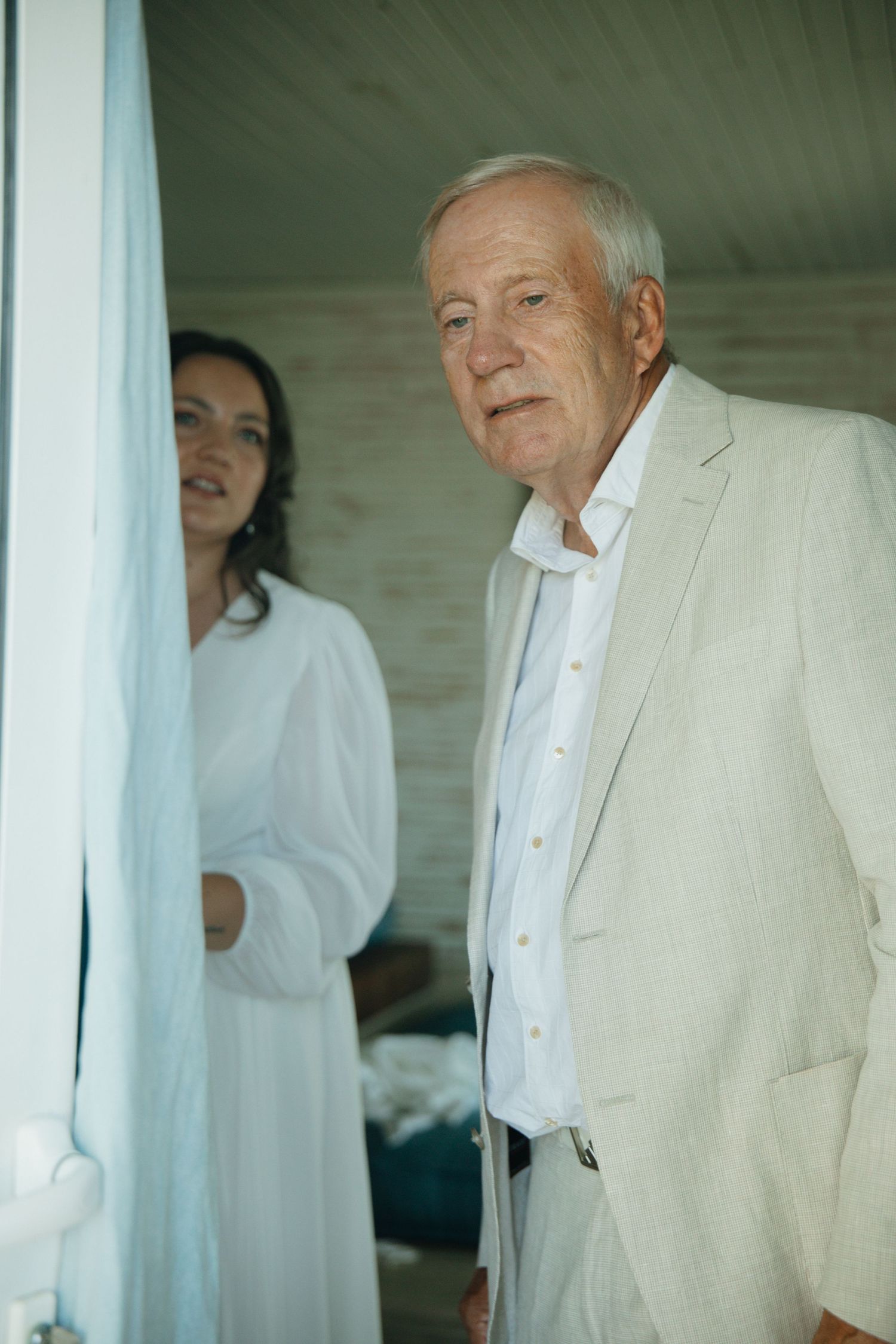 Elderly man in beige suit standing in doorway with woman in white dress behind him.