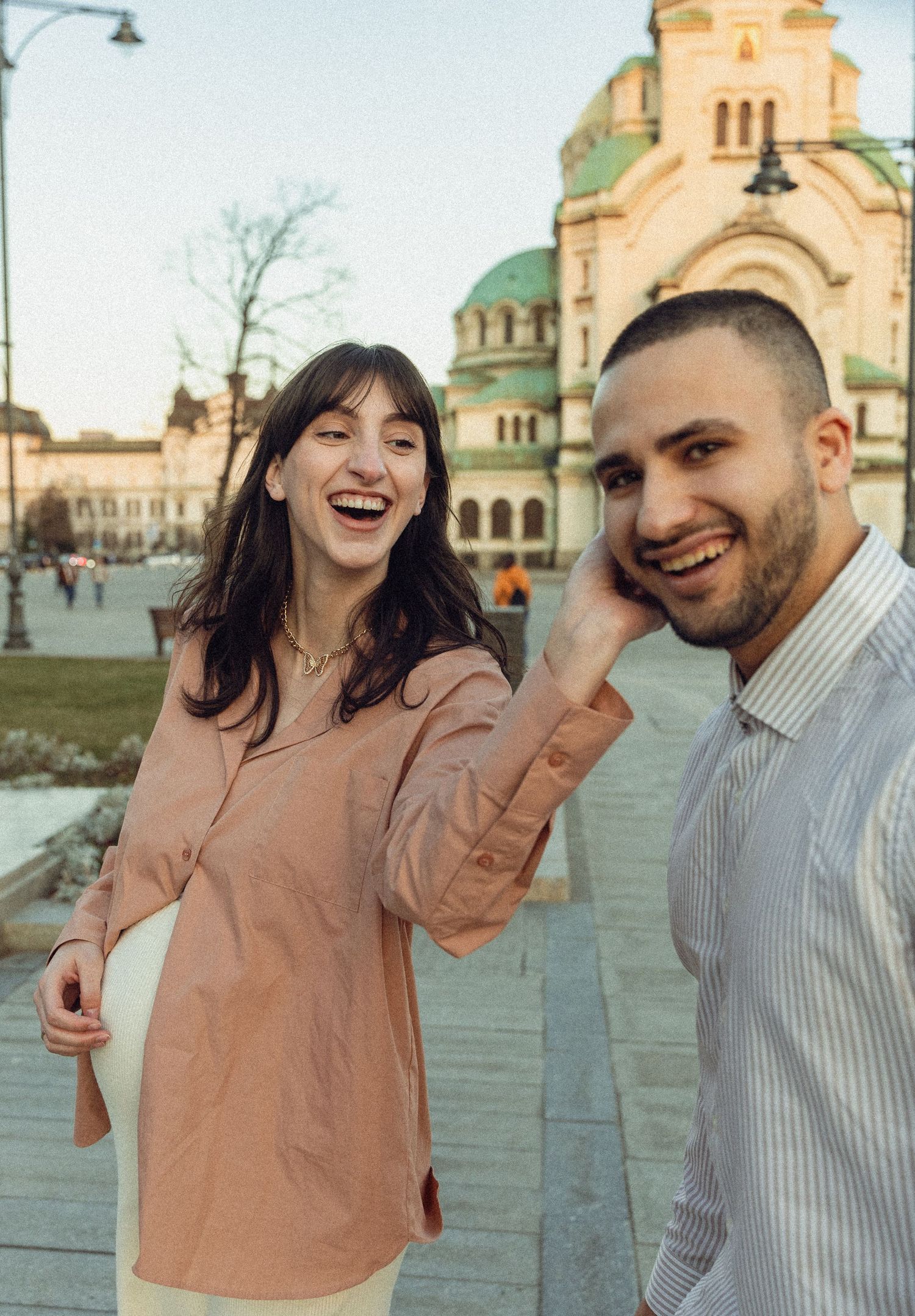 A laughing couple poses outdoors near a historic domed cathedral in Sofia, Bulgaria on a sunny day.