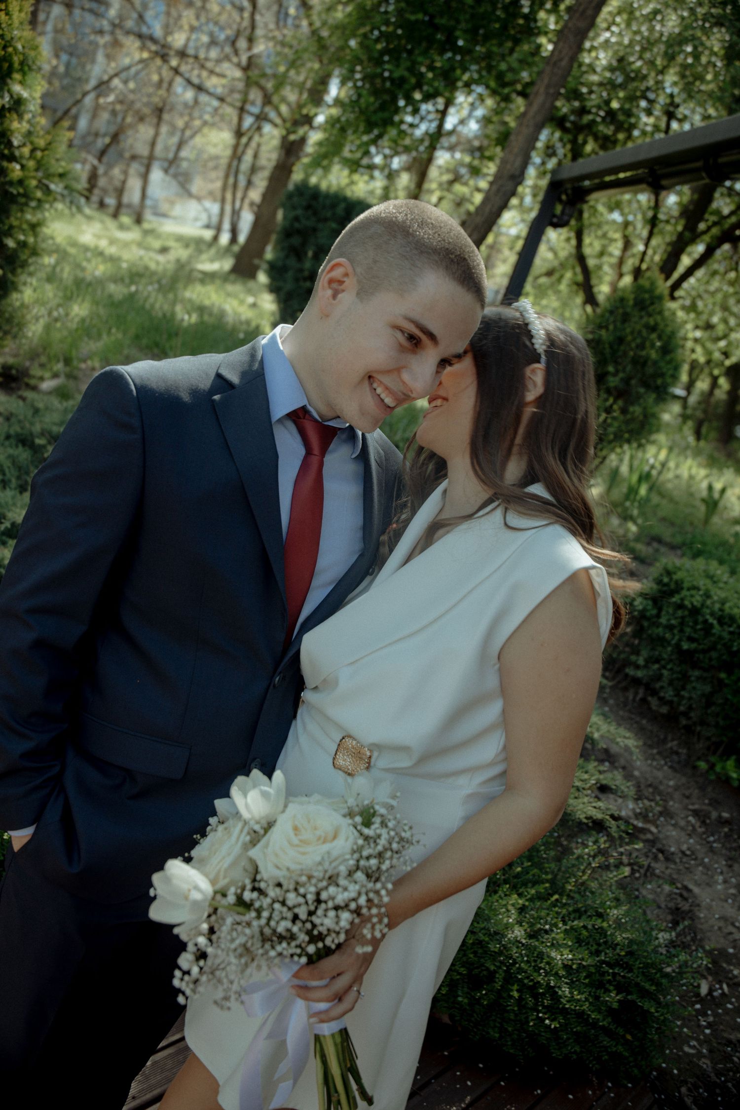 Smiling groom in navy suit and red tie leans toward bride in white dress holding white rose bouquet in a sunny garden.