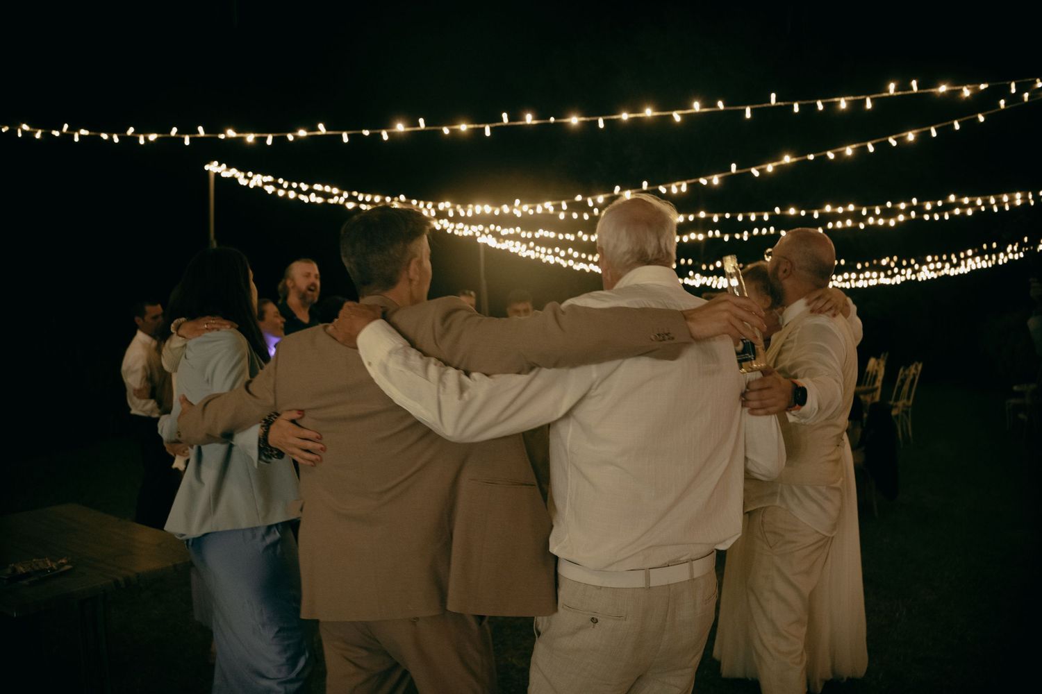 Group of men embracing under string lights at an outdoor evening celebration or wedding reception.