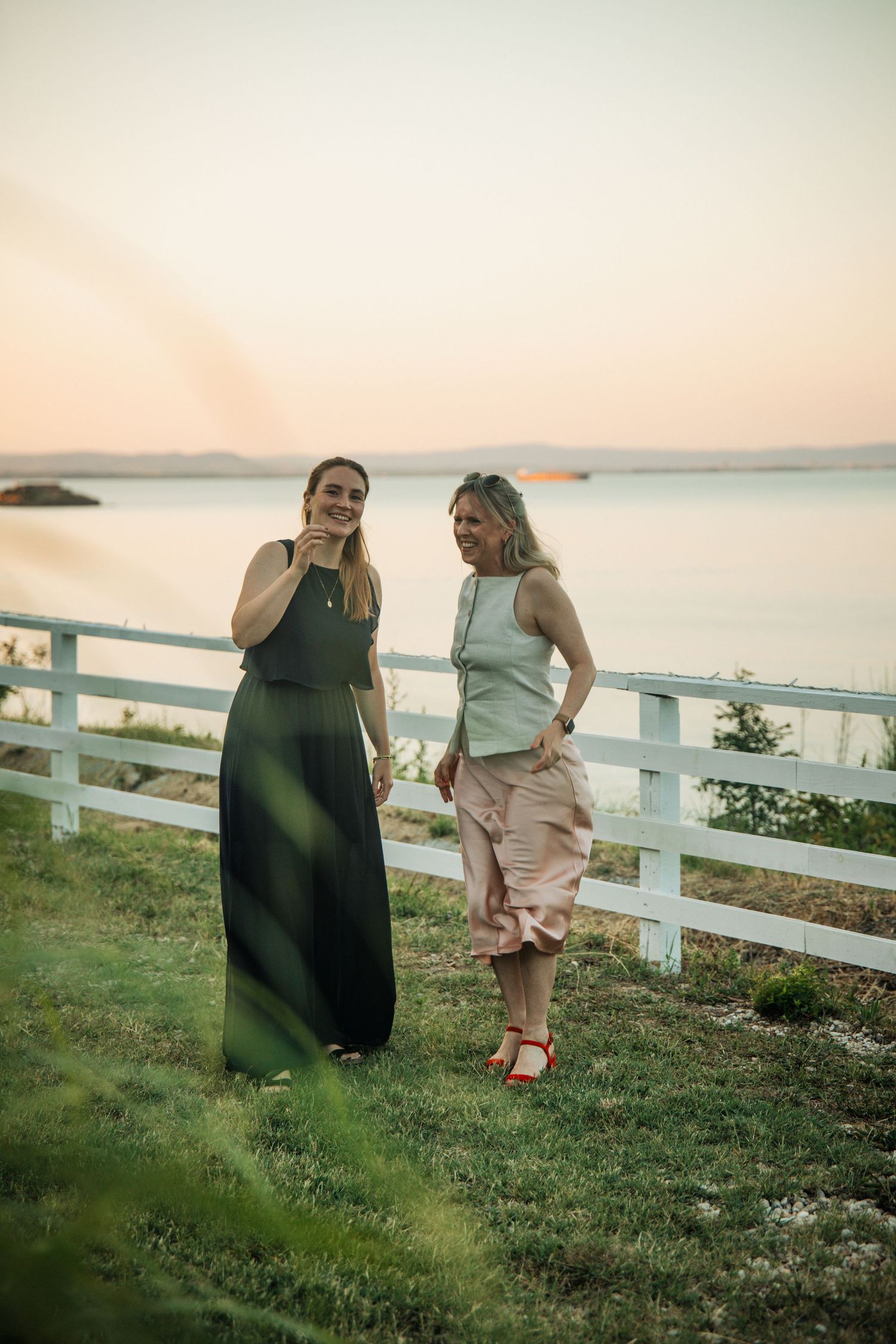 Two women in elegant dresses stand by white fence at sunset overlooking serene waterfront.
