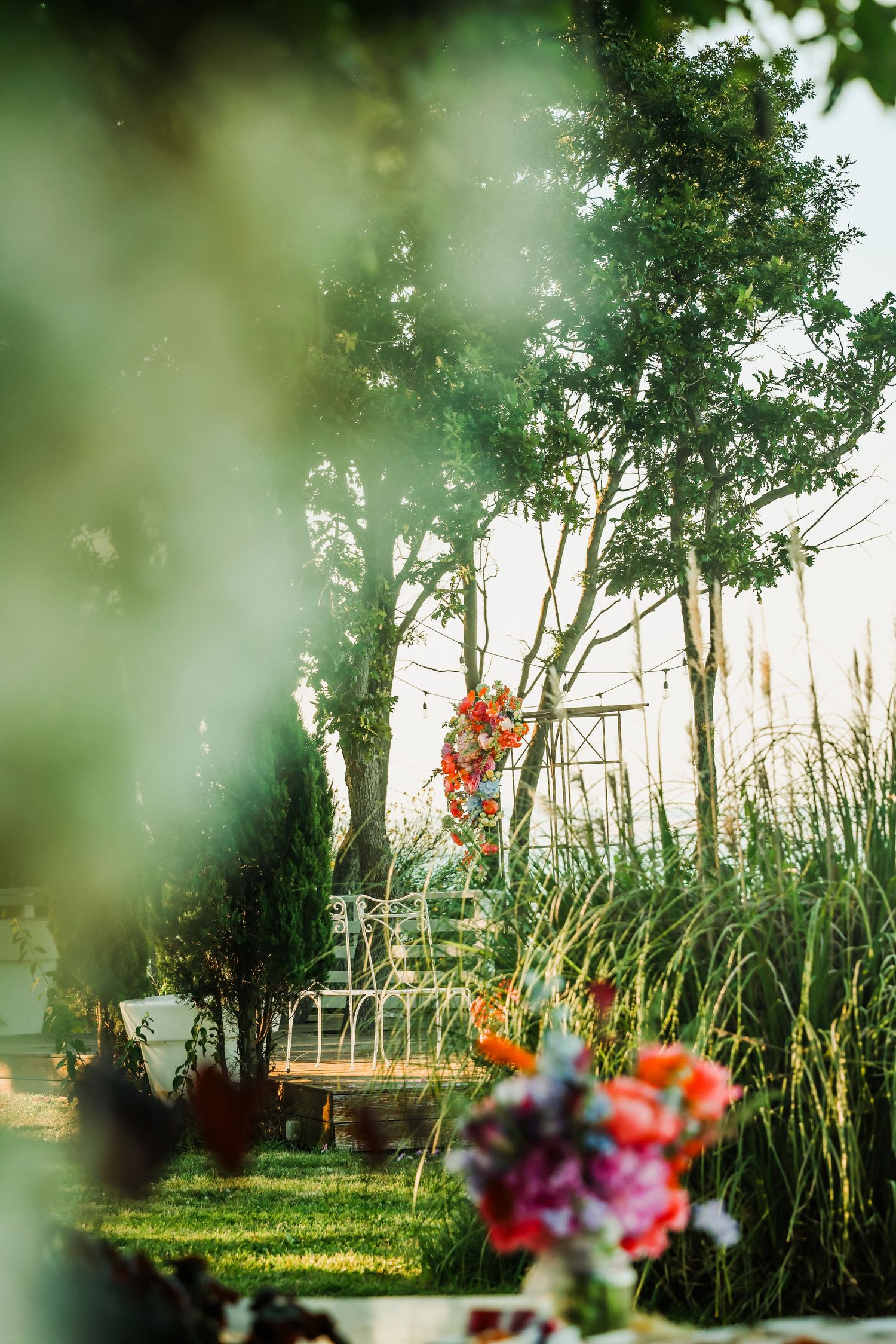Lush garden wedding venue with colorful floral arrangements, white bench, and tall trees bathed in warm golden sunlight.