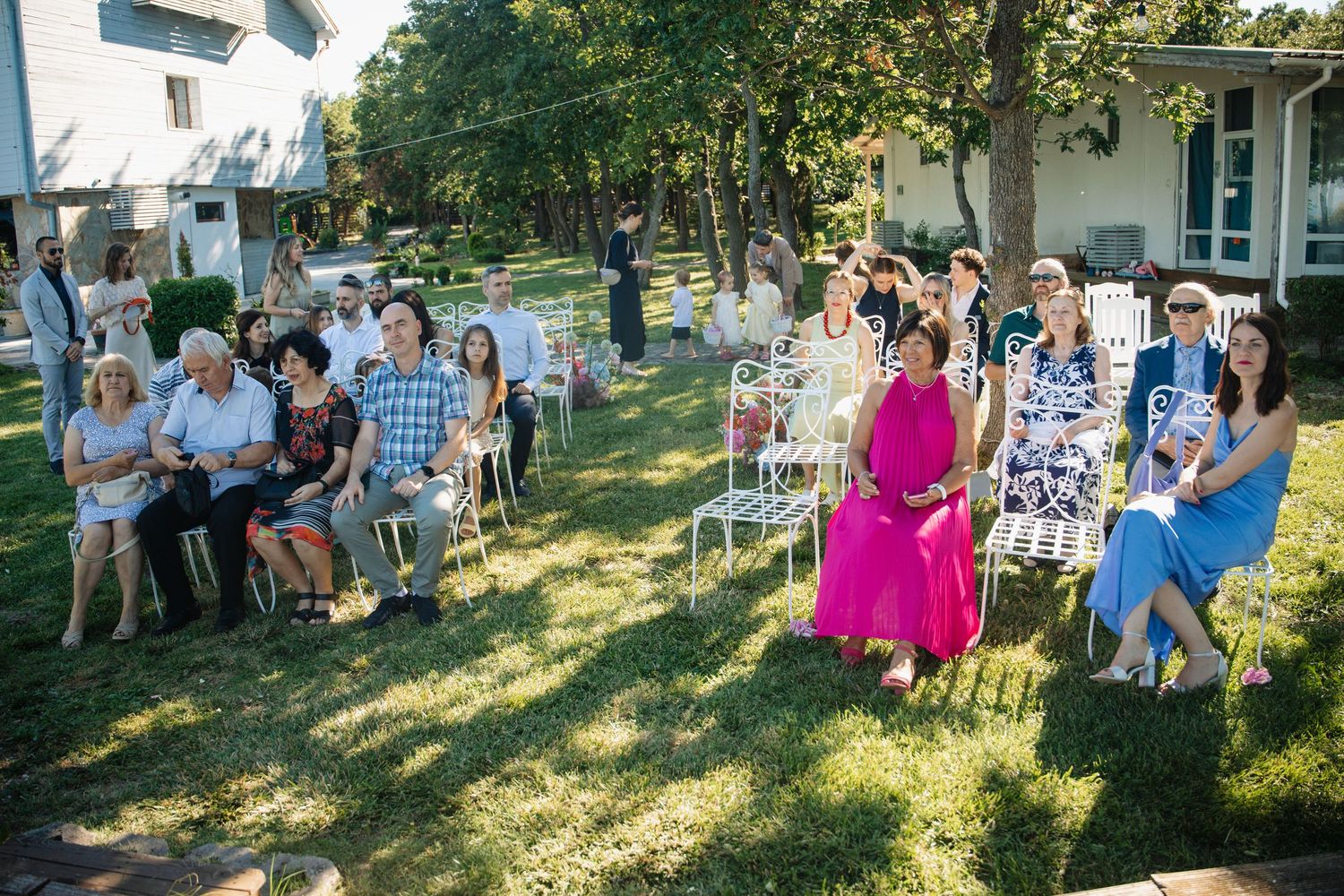 Guests seated on white chairs at an outdoor wedding ceremony on a sunny green lawn.