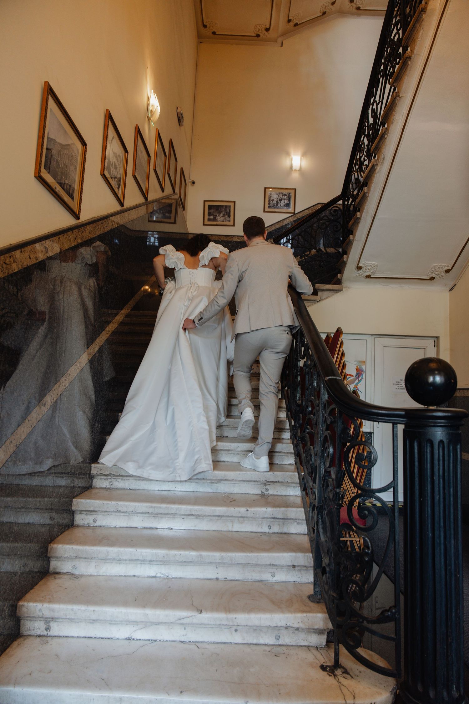 Bride and groom walking up elegant marble staircase with ornate iron railing in a classic wedding venue.